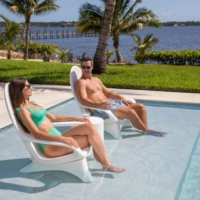 A man and woman relaxing by a pool, sitting on white lounge chairs with a lake view in the background.