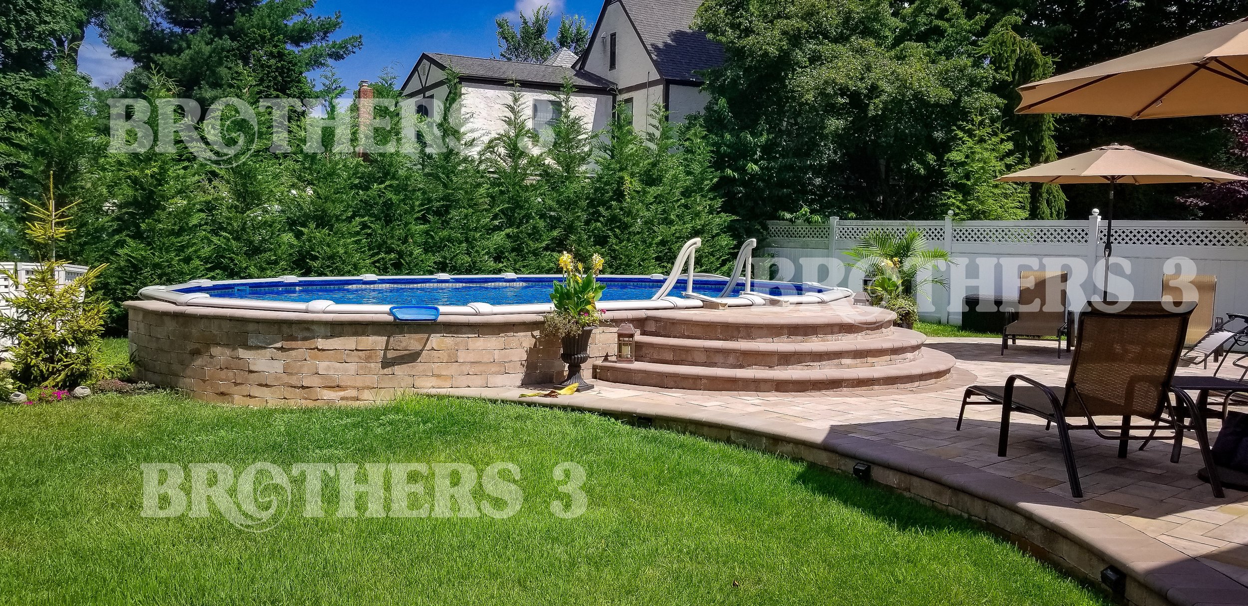 Backyard with an above-ground pool, brick steps, patio chairs with umbrellas, and a white fence, surrounded by trees and a house in the background.