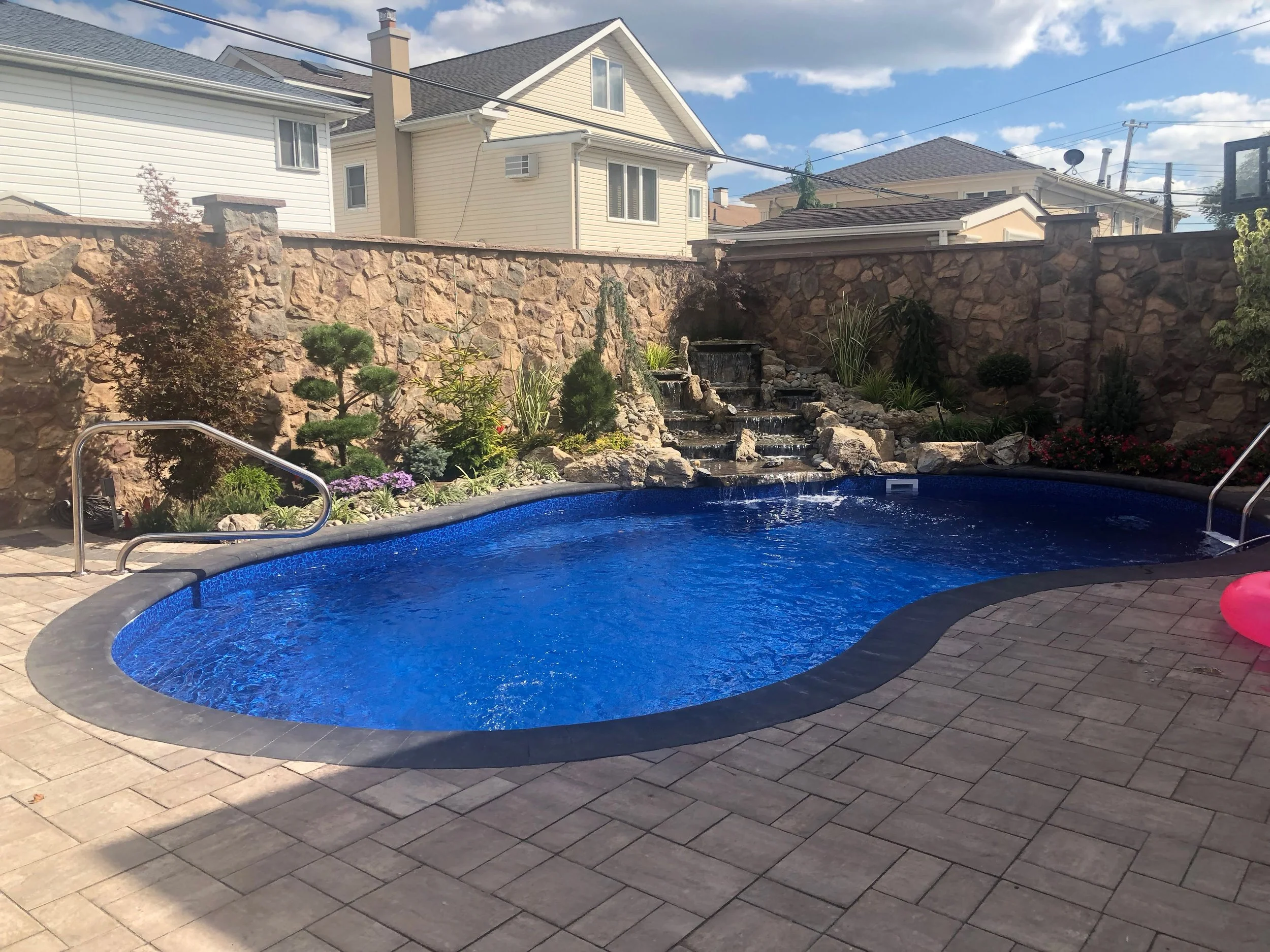 Backyard with in-ground swimming pool, waterfall feature, stone wall, landscaped plants, and patio with pavers.