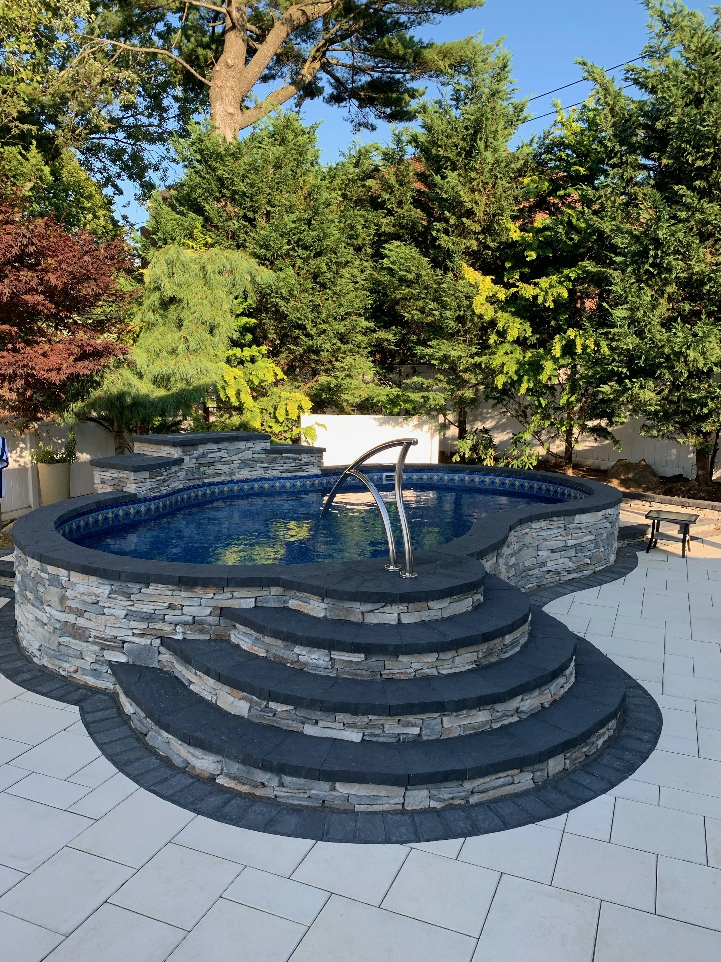 An outdoor backyard pool with curved stone steps leading into the pool, surrounded by a patio with white tiles, tall trees in the background, and a clear blue sky.
