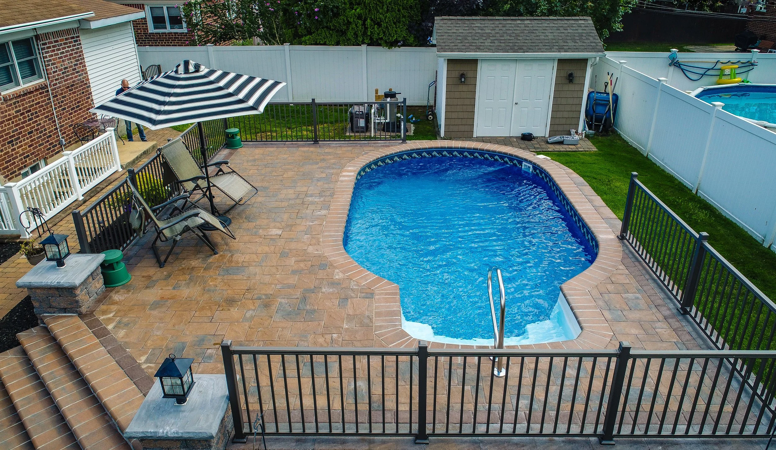 A backyard with a swimming pool, patio with outdoor chairs and umbrella, white fence, small lawn area, and storage shed.
