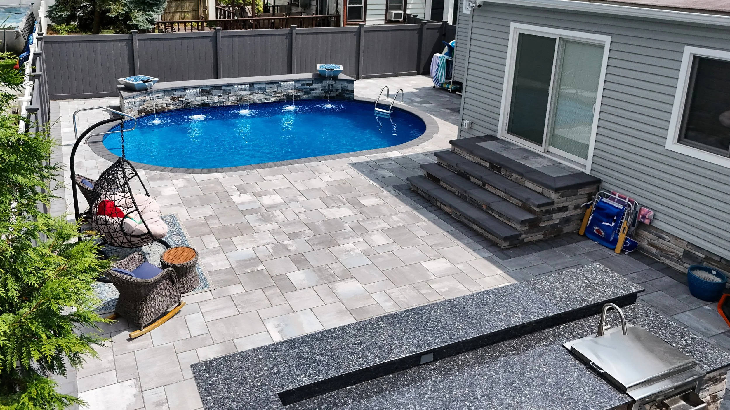 Backyard with a small oval swimming pool, a gray fence, steps leading to a sliding glass door, outdoor furniture, and a shaded area with a hanging chair. There is also a built-in outdoor grill and various pool accessories.