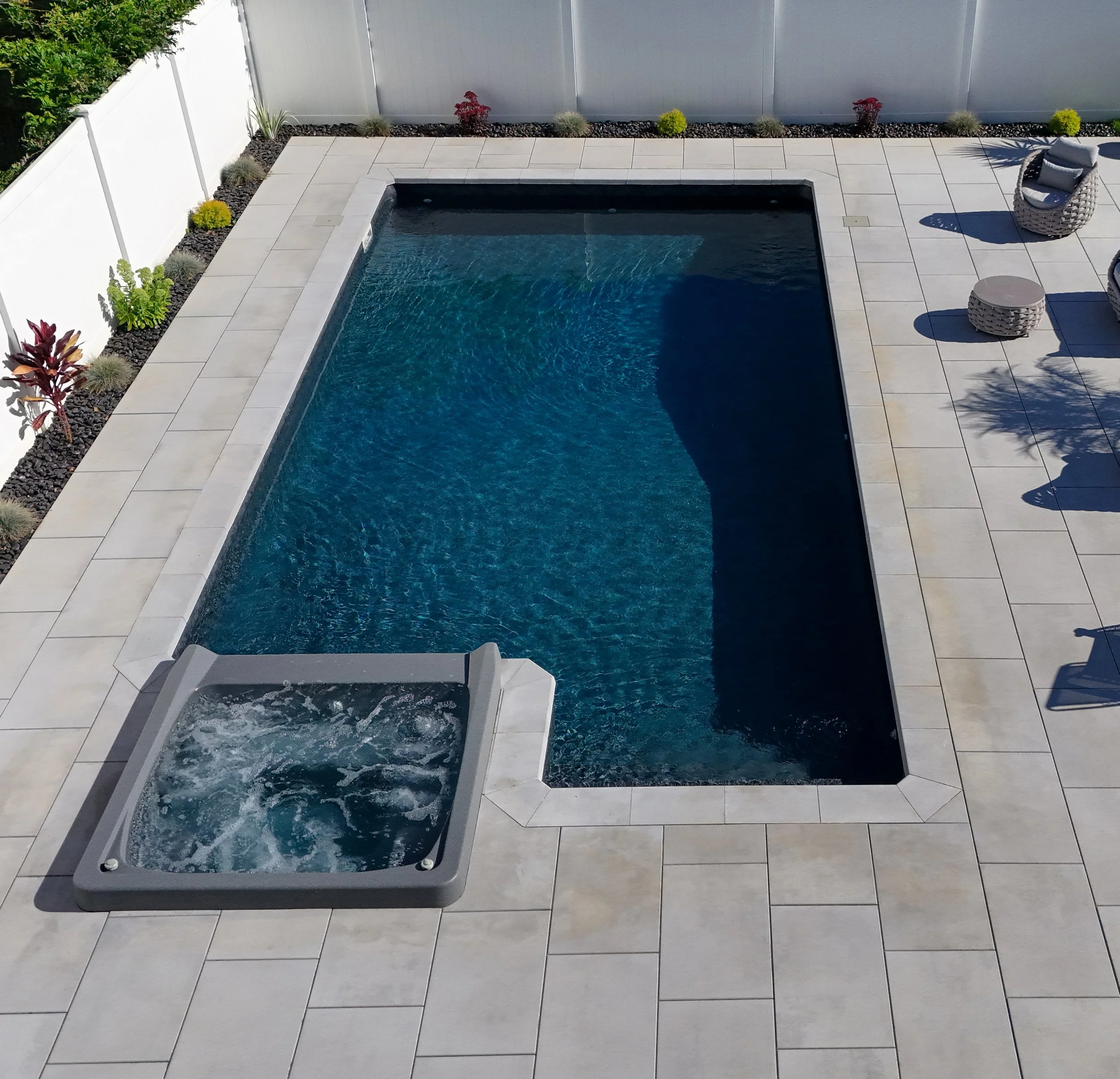 A rectangular swimming pool with a hot tub attached in a backyard with a tiled patio and a white fence. There are outdoor chairs, plants, and shadows cast by the chairs and plants.