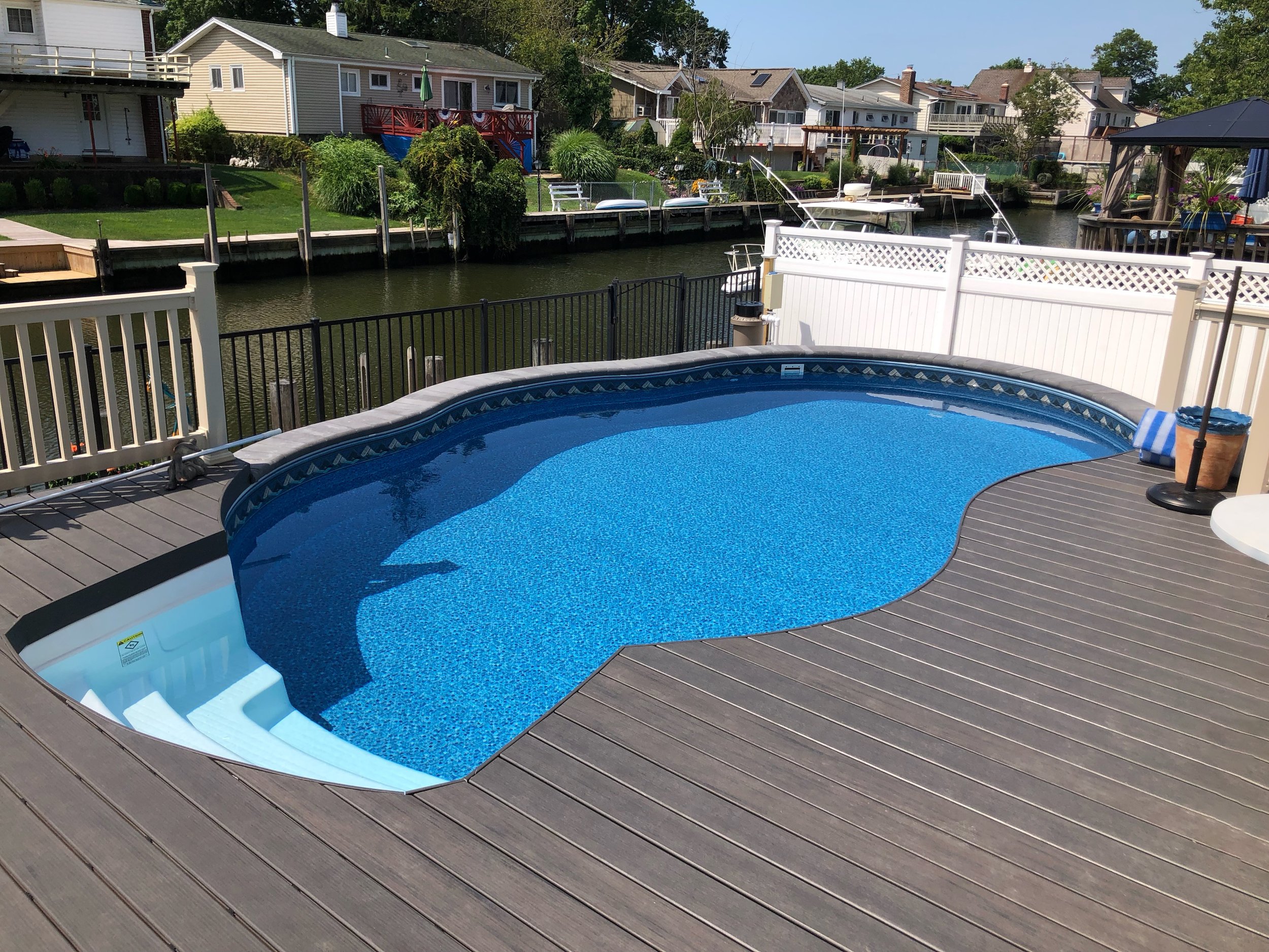 In-ground swimming pool on a wooden deck beside a canal with boats, houses, and greenery in the background.