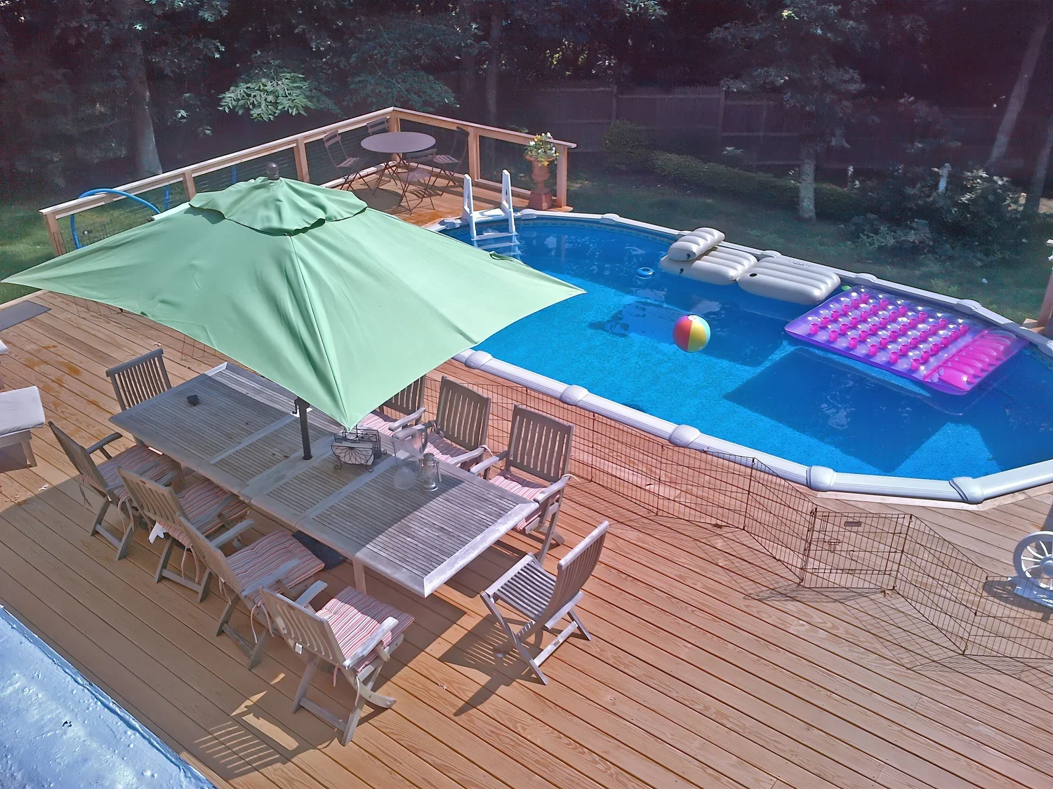 A backyard pool area with a wooden deck, a large green umbrella, a dining table with chairs, an inflatable pool with a float, a beach ball, and a pink geometric-patterned float, surrounded by a wooden fence and trees.