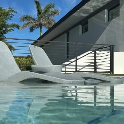 Modern poolside lounge chairs near a swimming pool with a house and palm trees in the background.