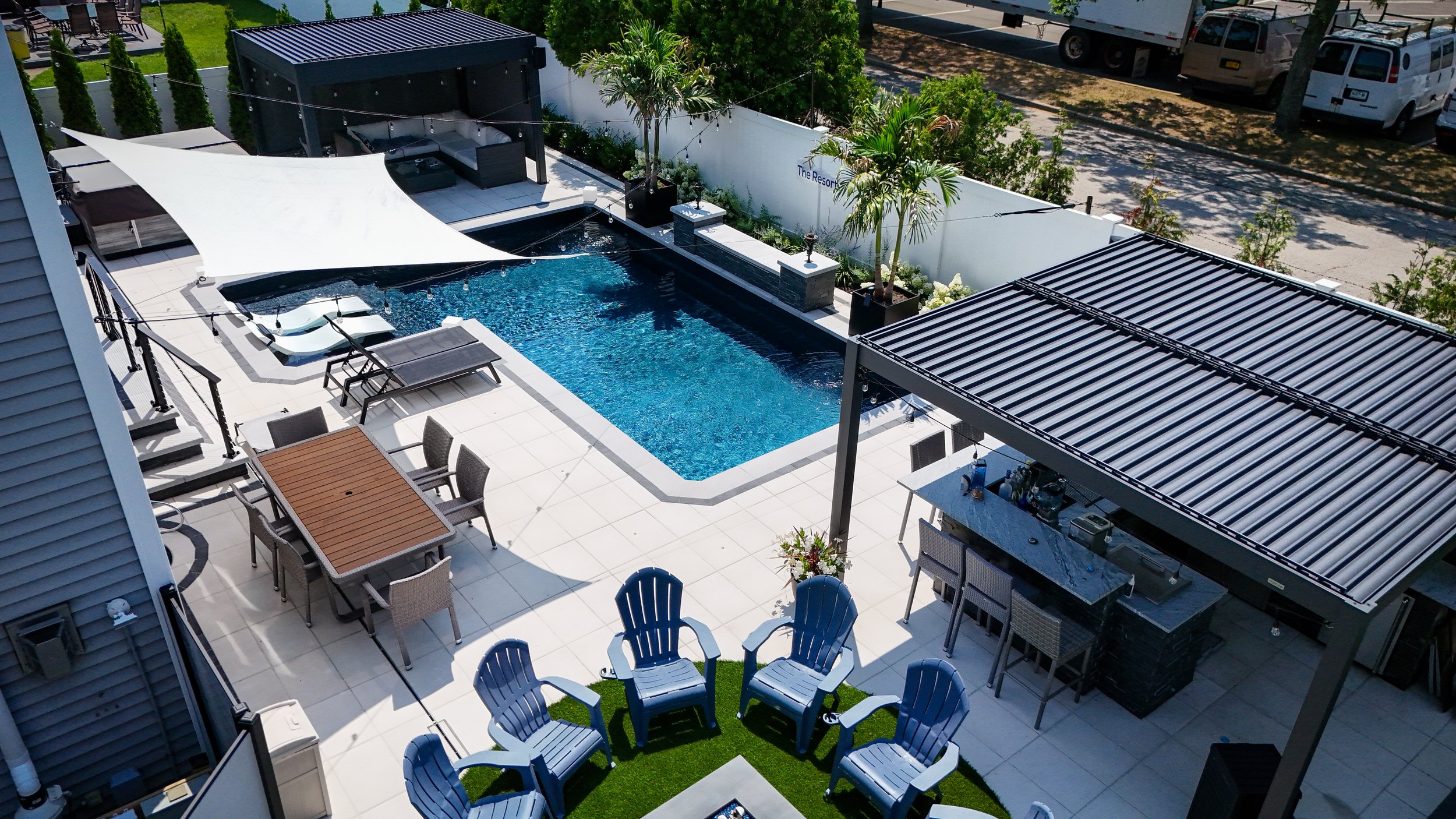 A rooftop outdoor area with a swimming pool, lounge chairs, a shaded bar area, and a seating area with blue Adirondack chairs. There are trees, potted plants, and parked vehicles in the background.