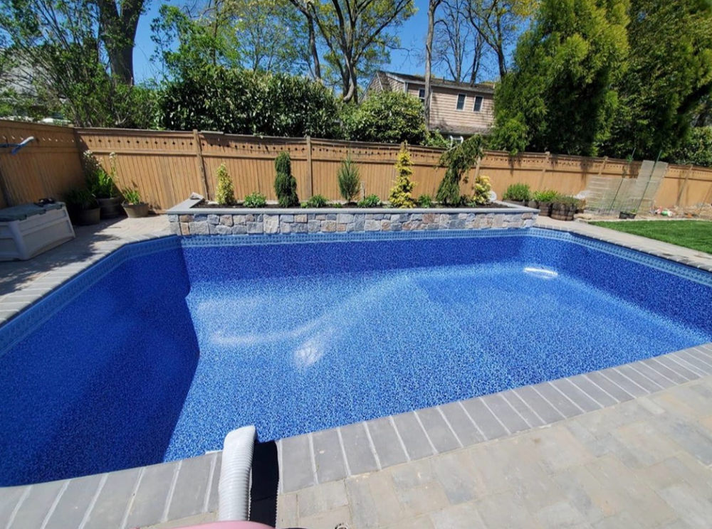 Residential backyard swimming pool with a stone border, surrounded by a brick patio, a wooden fence, and landscaped plants, with trees and a house in the background.
