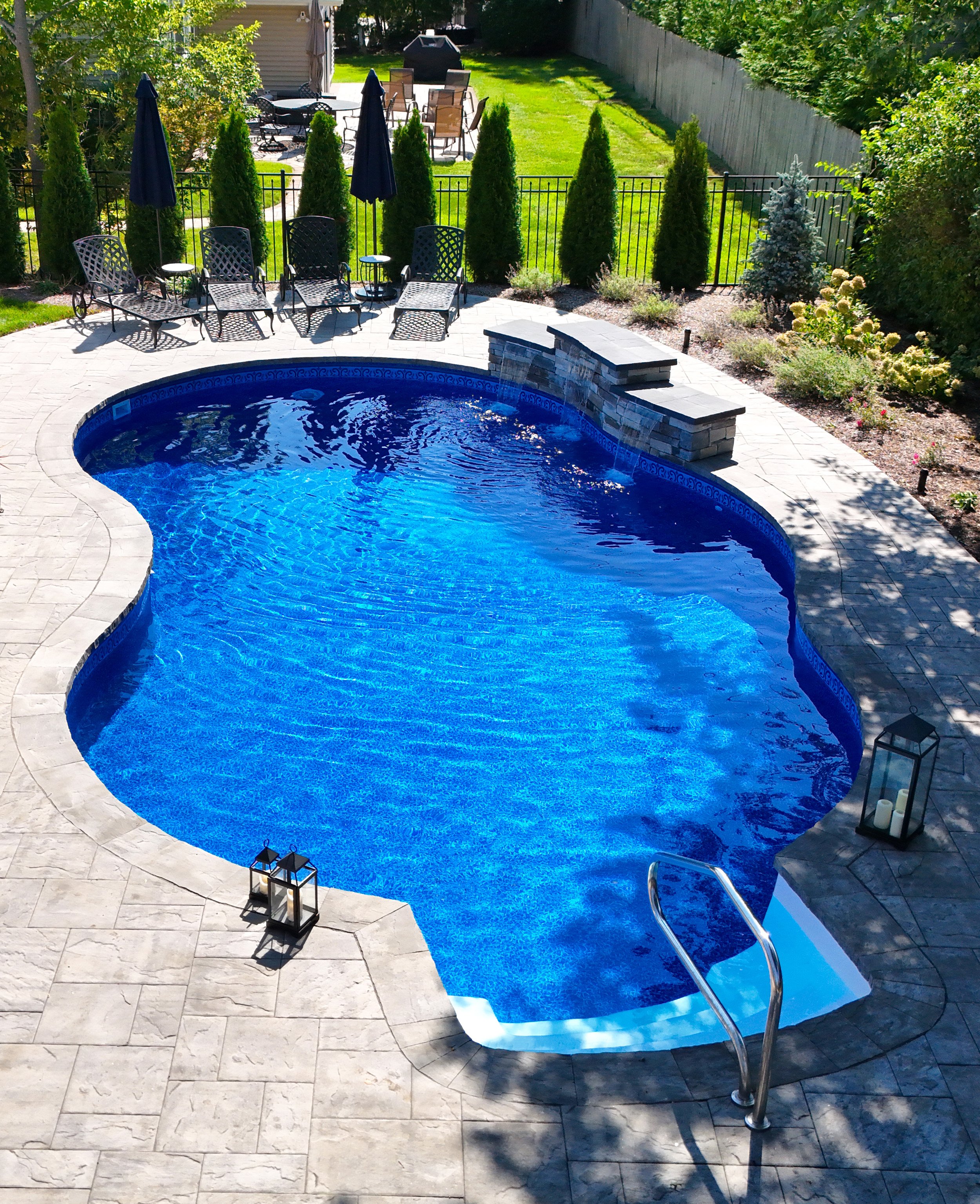 A backyard with a swimming pool, patio furniture, umbrellas, a fenced yard, and landscaped plants and trees.