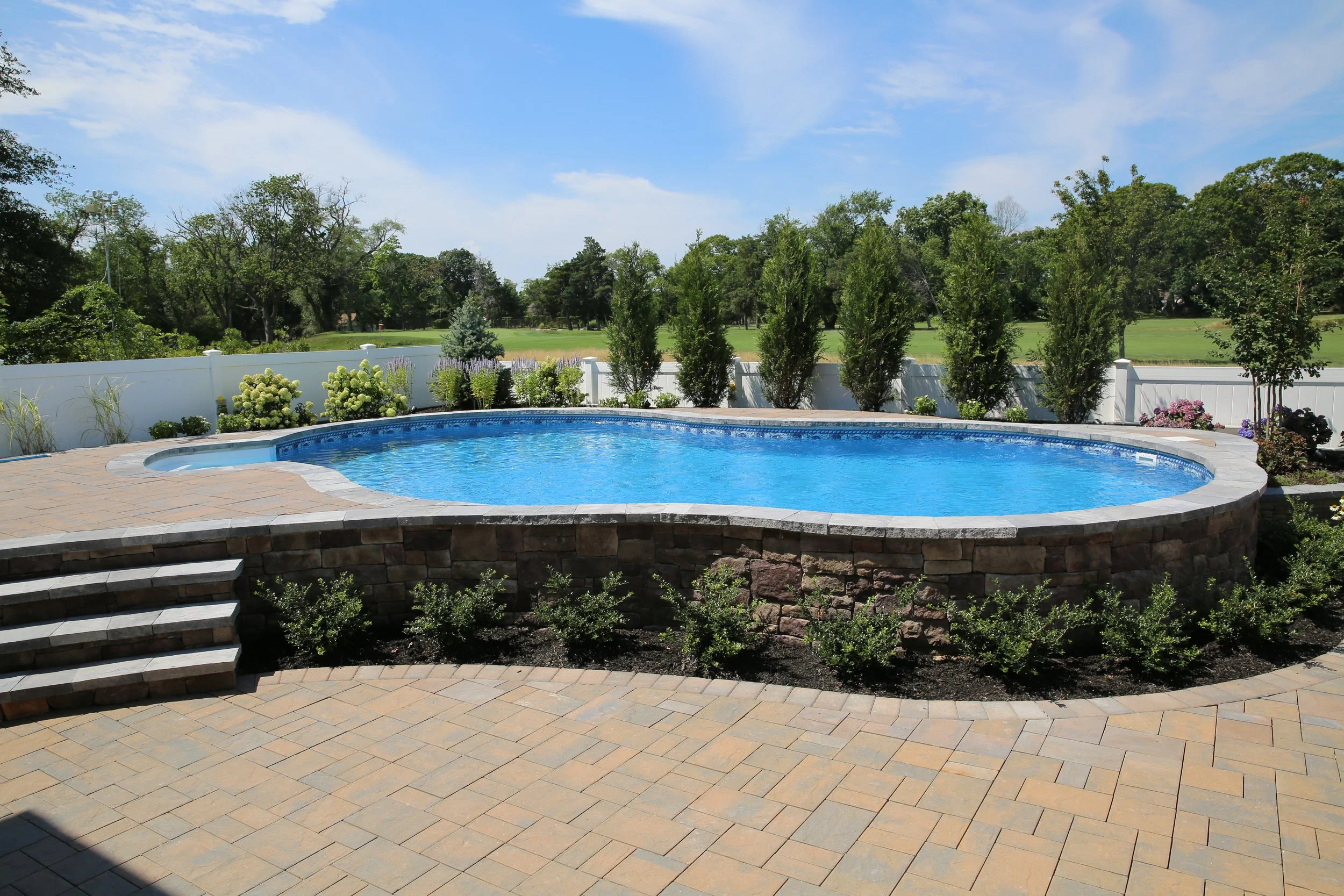A backyard with a blue in-ground swimming pool, edged with stone, surrounded by a paved patio and landscaped with plants and shrubs, enclosed by a white fence and green trees under a partly cloudy sky.