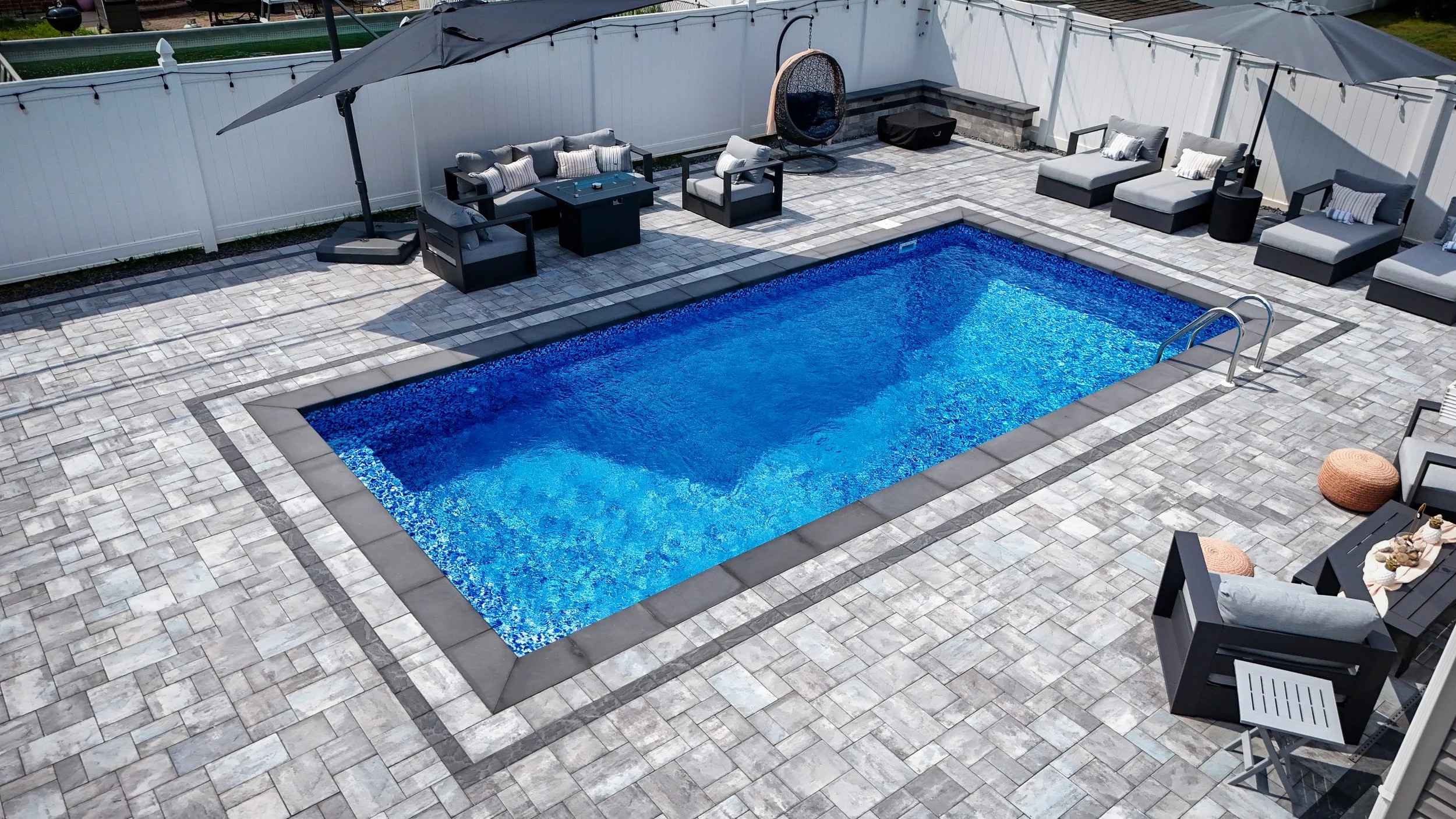 A backyard with a rectangular blue swimming pool surrounded by gray paving stones. There are outdoor lounge chairs, a covered patio umbrella, a hanging chair, and various outdoor furniture and accessories around the pool.