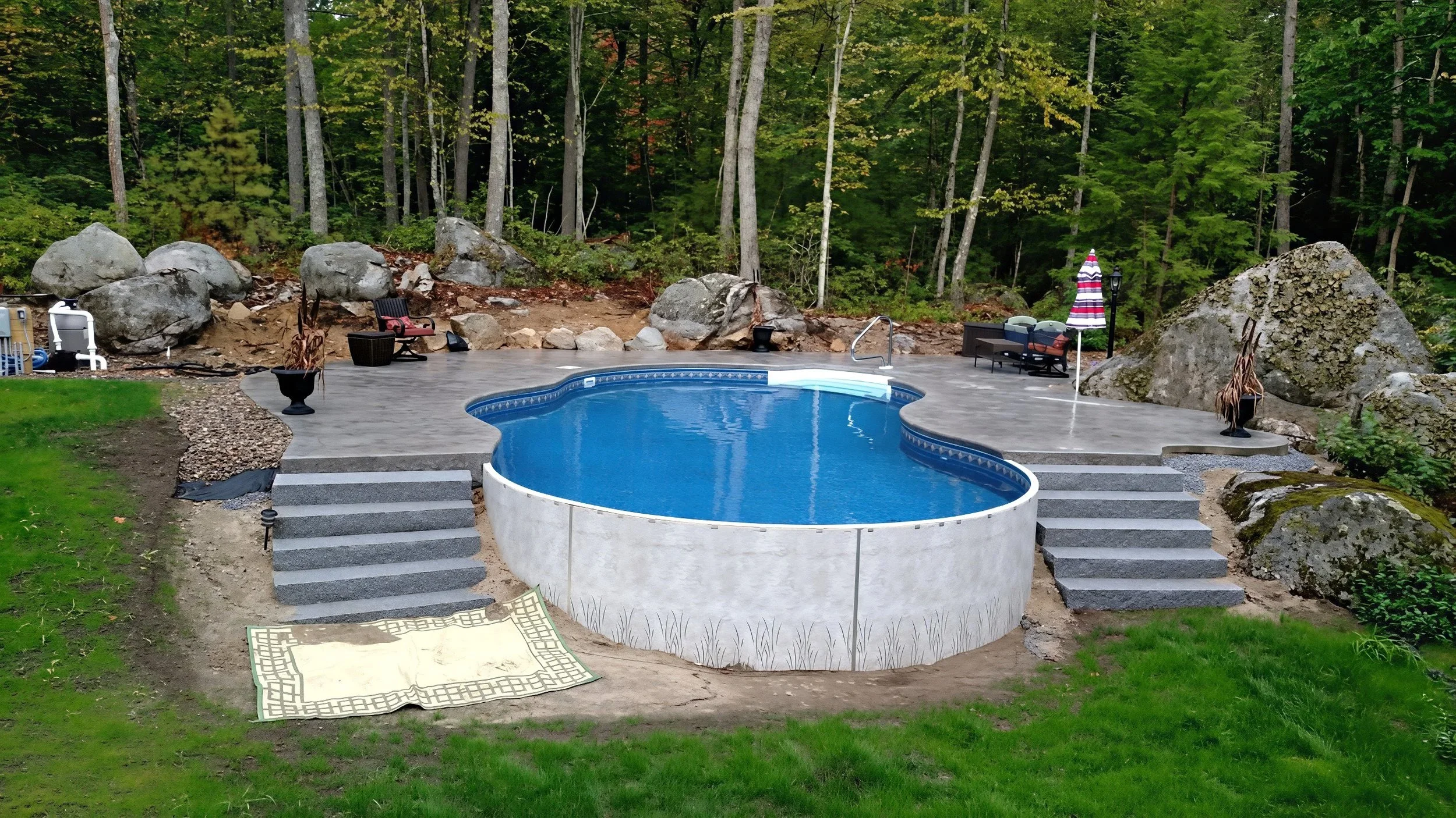 A backyard with a heart-shaped swimming pool surrounded by a stone patio, stairs, and large rocks. There are chairs, a table with an umbrella, and a forested background.