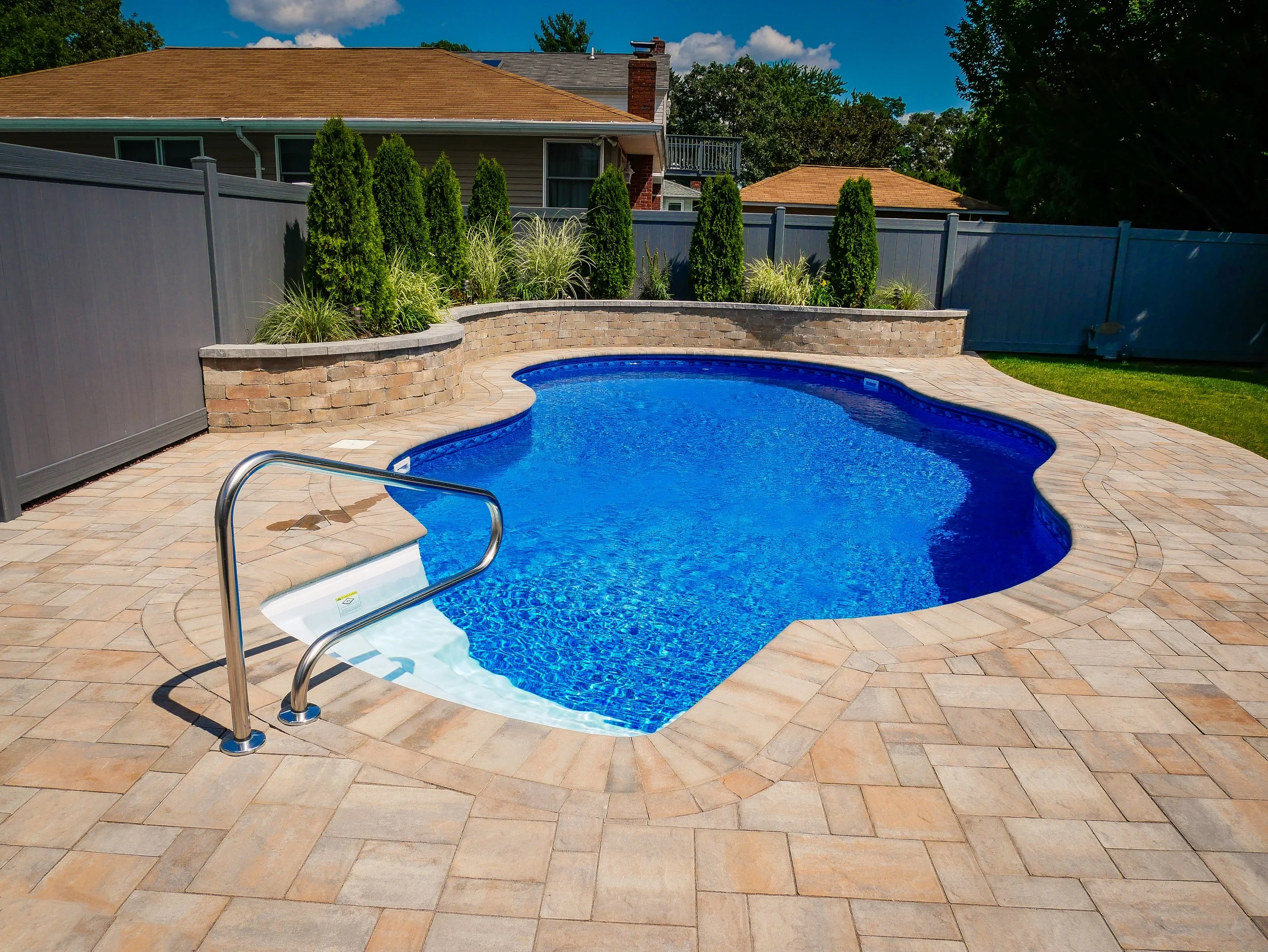 Backyard with a kidney-shaped swimming pool, surrounded by a stone deck, bordered by a gray fence, with tall evergreen shrubs and plants on a brick wall in the background, and houses beyond that.
