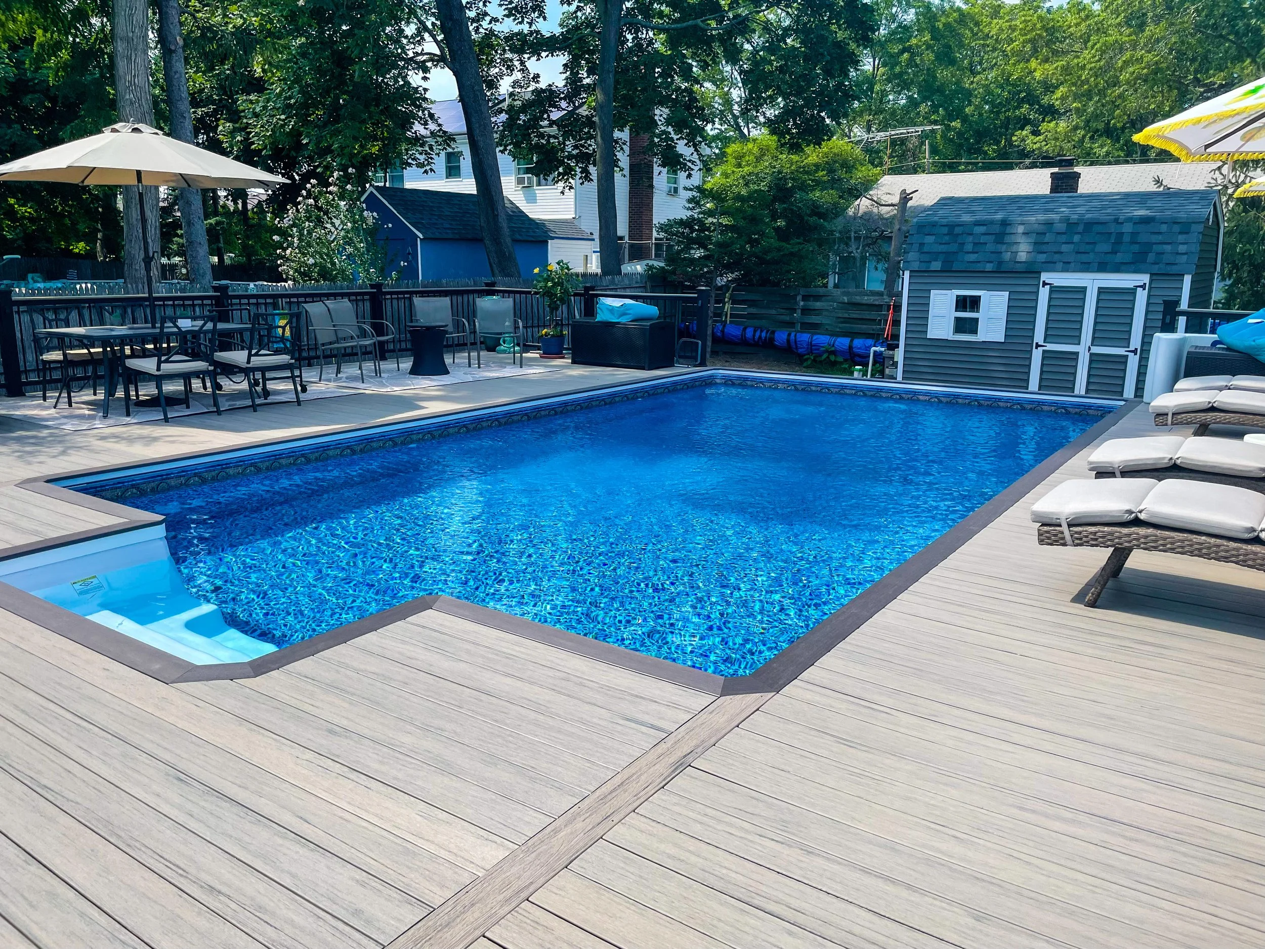 An outdoor backyard scene featuring a blue swimming pool with a small attached hot tub, surrounded by a light-colored wooden deck with lounge chairs, a patio table with chairs under an umbrella, and a small blue shed in the background, with trees and neighboring houses visible.
