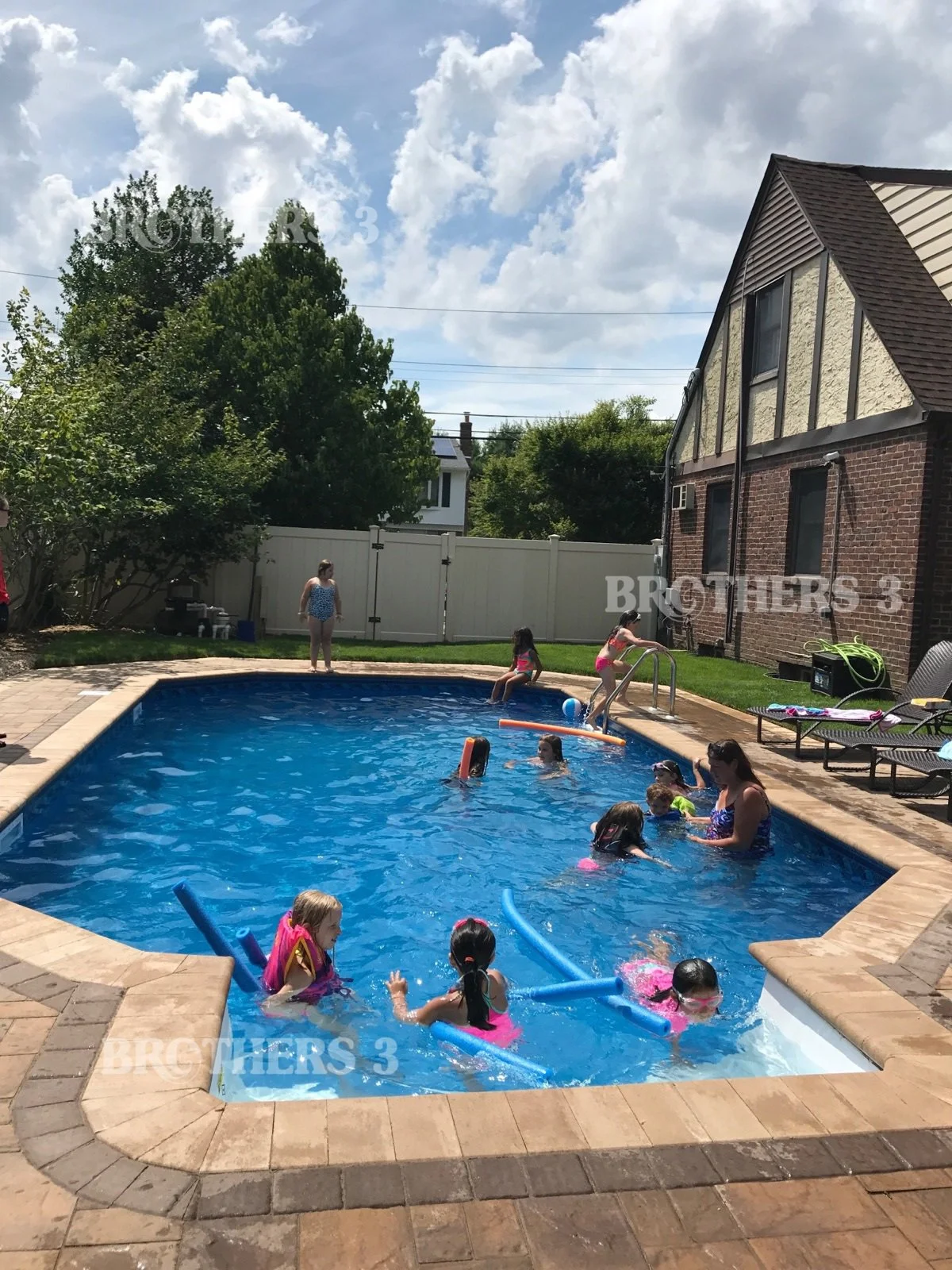 Children swimming and playing in a backyard swimming pool on a sunny day, with adults supervising, surrounded by a brick patio, green grass, trees, and a house with a steep roof and gable.