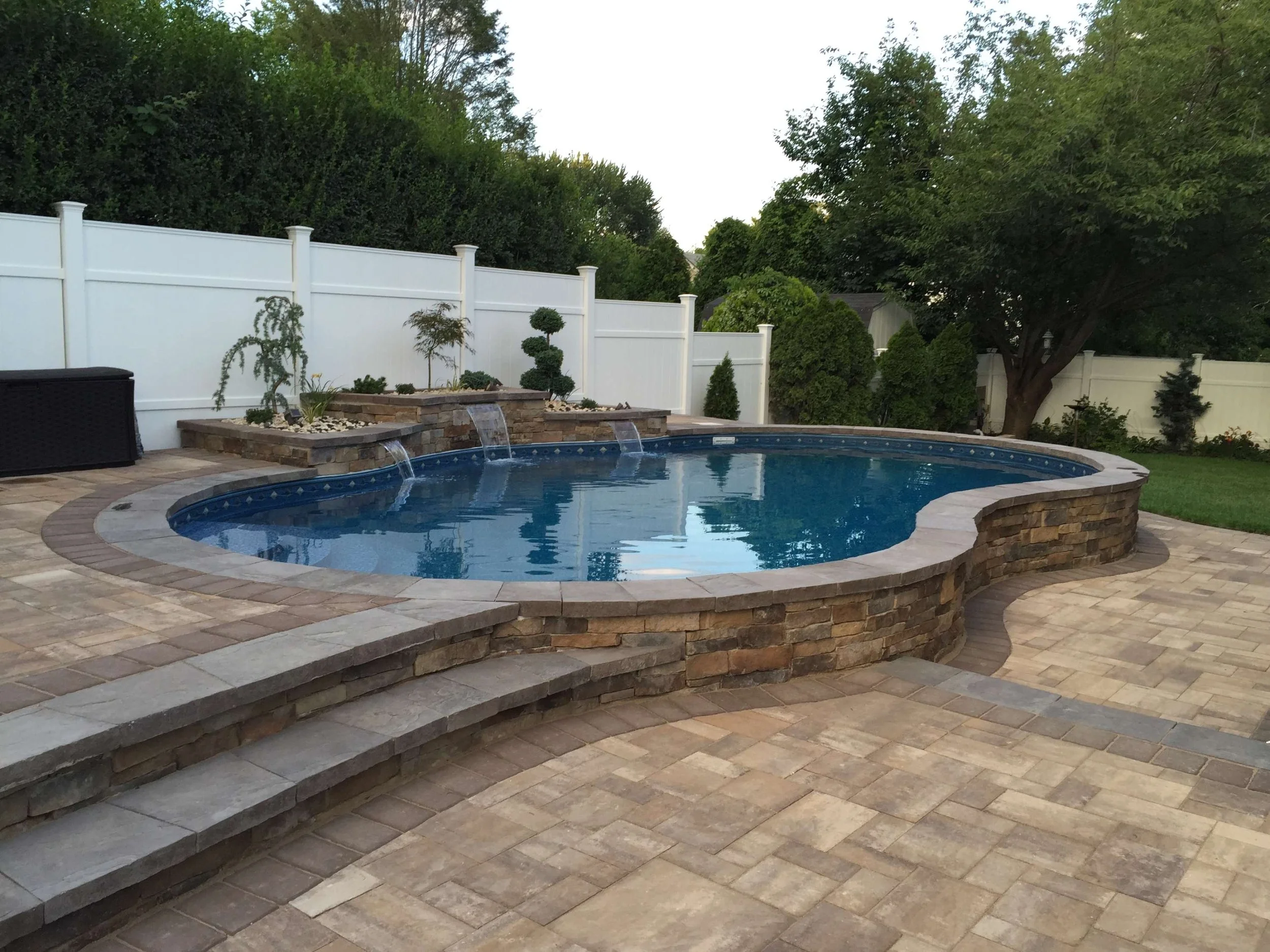 Residential backyard with a kidney-shaped swimming pool, stone patio, and a white fence. There are water features on a raised brick platform with small waterfall spouts, trees, and shrubs in the background.