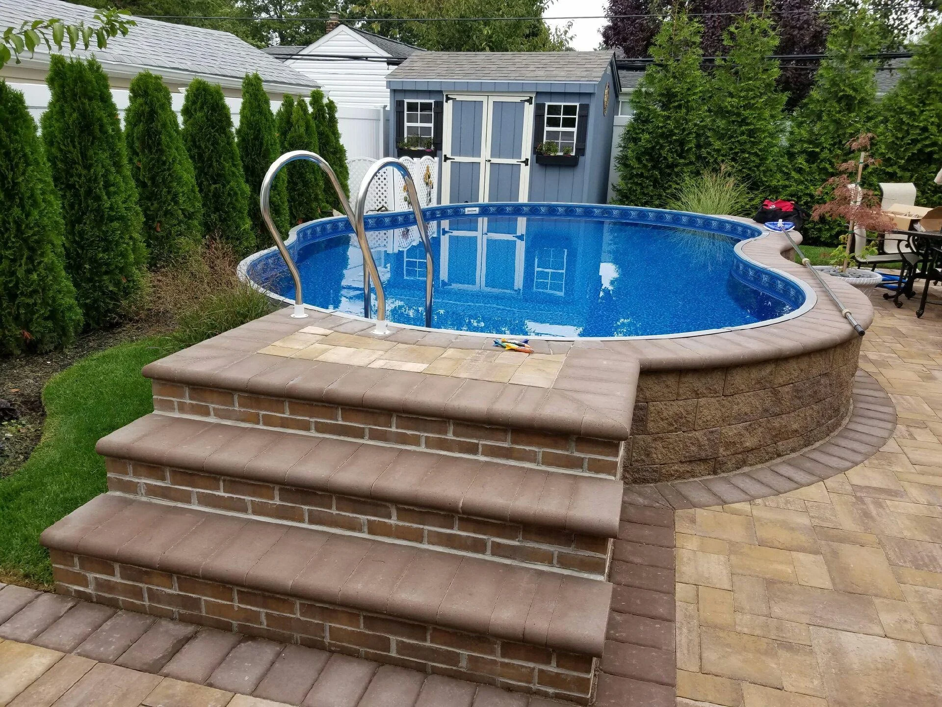 A backyard with a round above-ground swimming pool with blue water, metal ladder, brick steps, surrounding paver patio, green shrubbery, trees, a shed, and outdoor furniture.