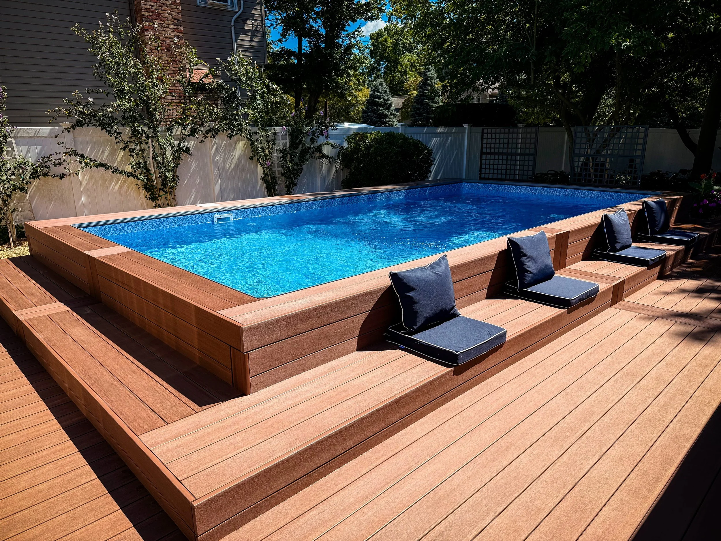 A backyard with a rectangular above-ground pool with a wooden deck and four black cushions with white trim on the edge.