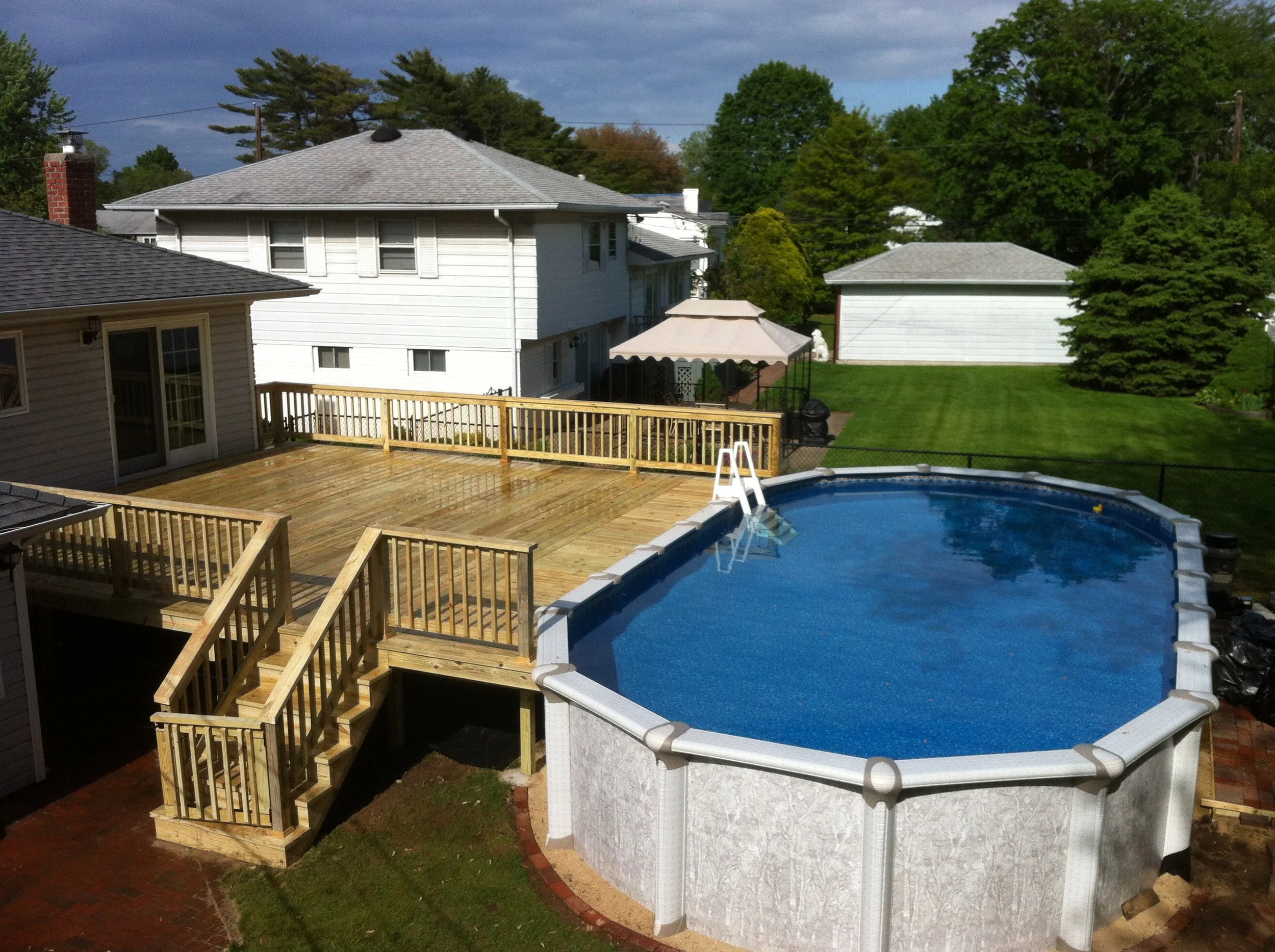 Backyard with above ground pool, wooden deck, and green lawn with trees.