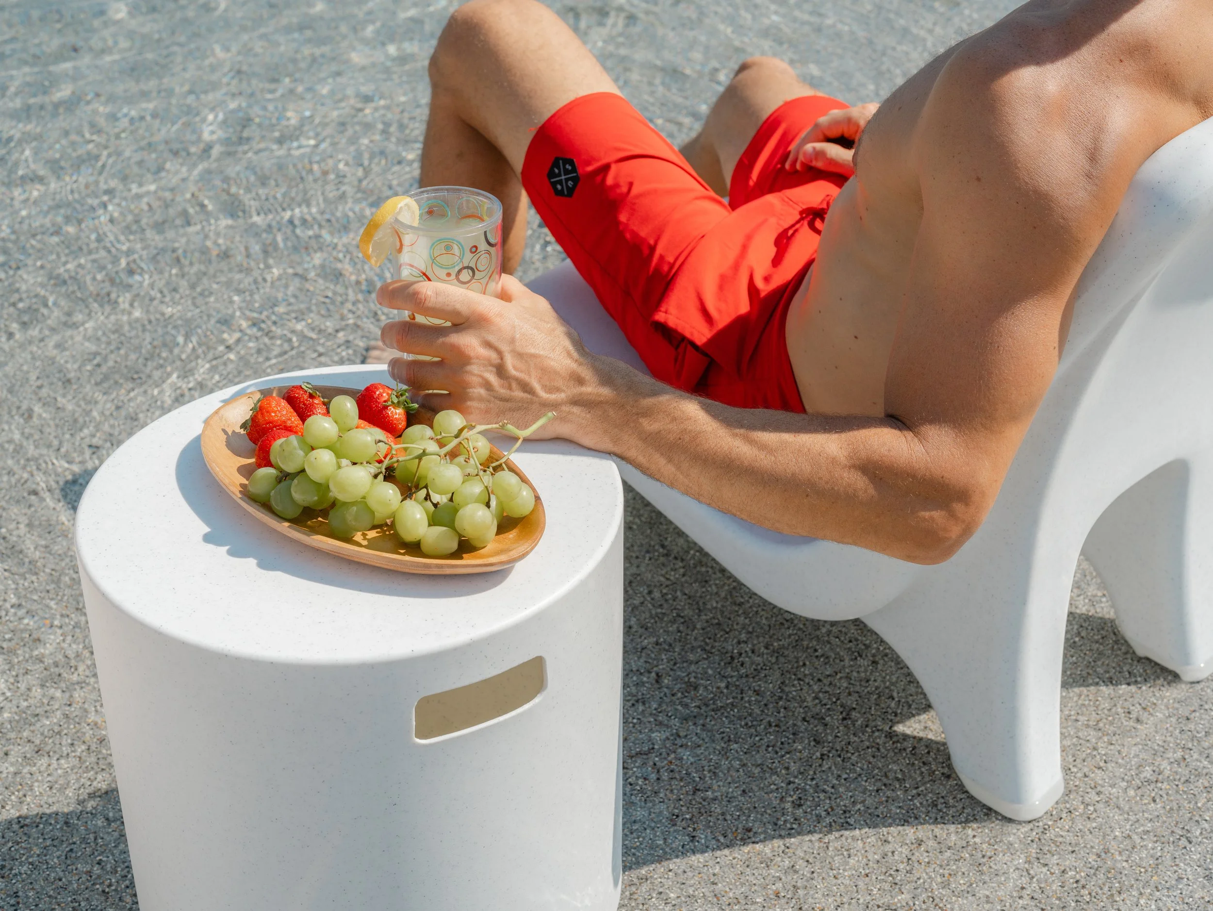 A man in red shorts relaxing on a white lounge chair on a sandy beach, holding a glass with a lemon slice, next to a plate of strawberries and green grapes on a small white table.