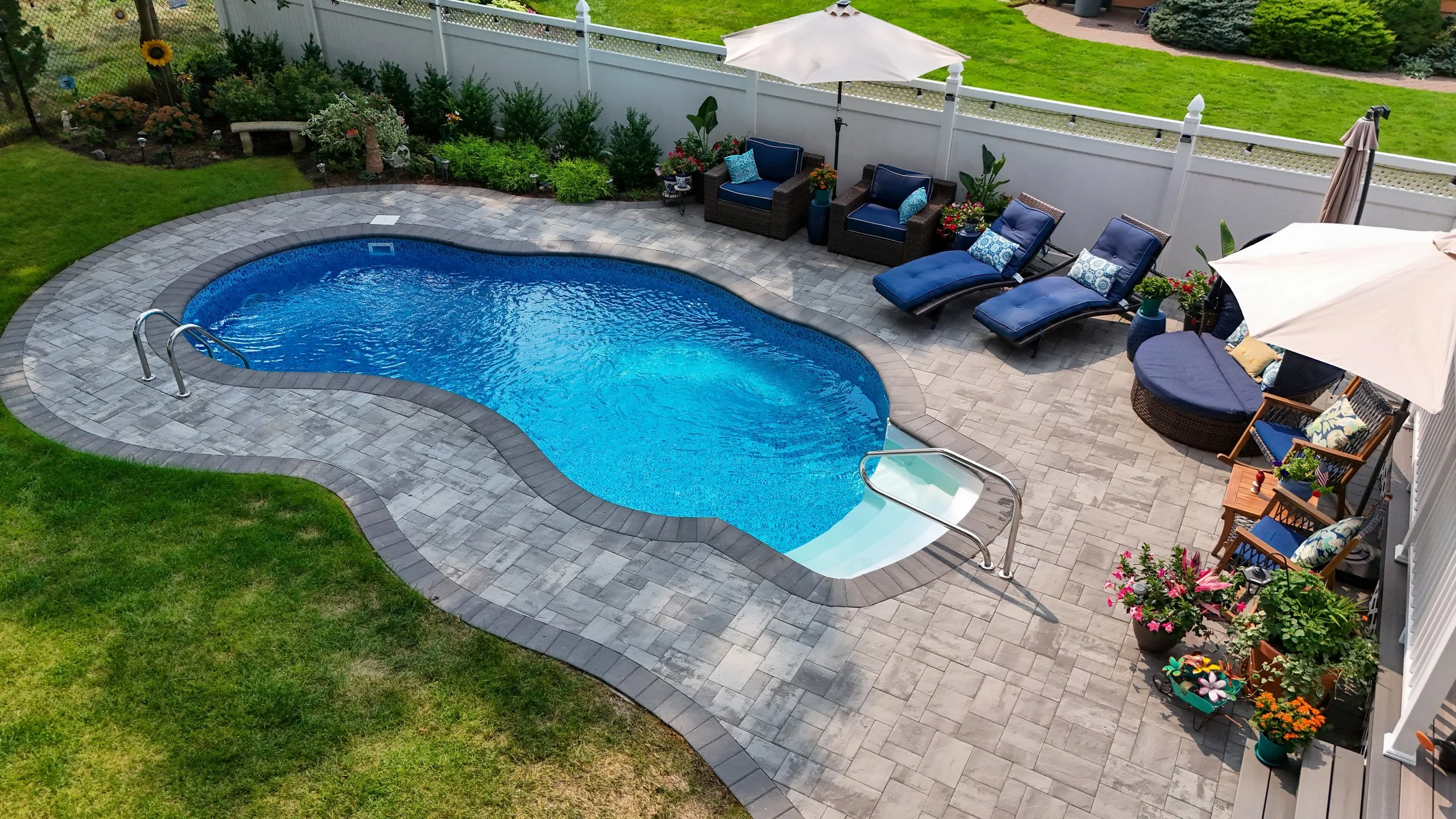 An outdoor backyard scene with a kidney-shaped swimming pool surrounded by a gray stone deck. There are sun loungers, chairs, umbrellas, and potted plants with flowers along a white fence, with a well-maintained lawn and garden area beyond.