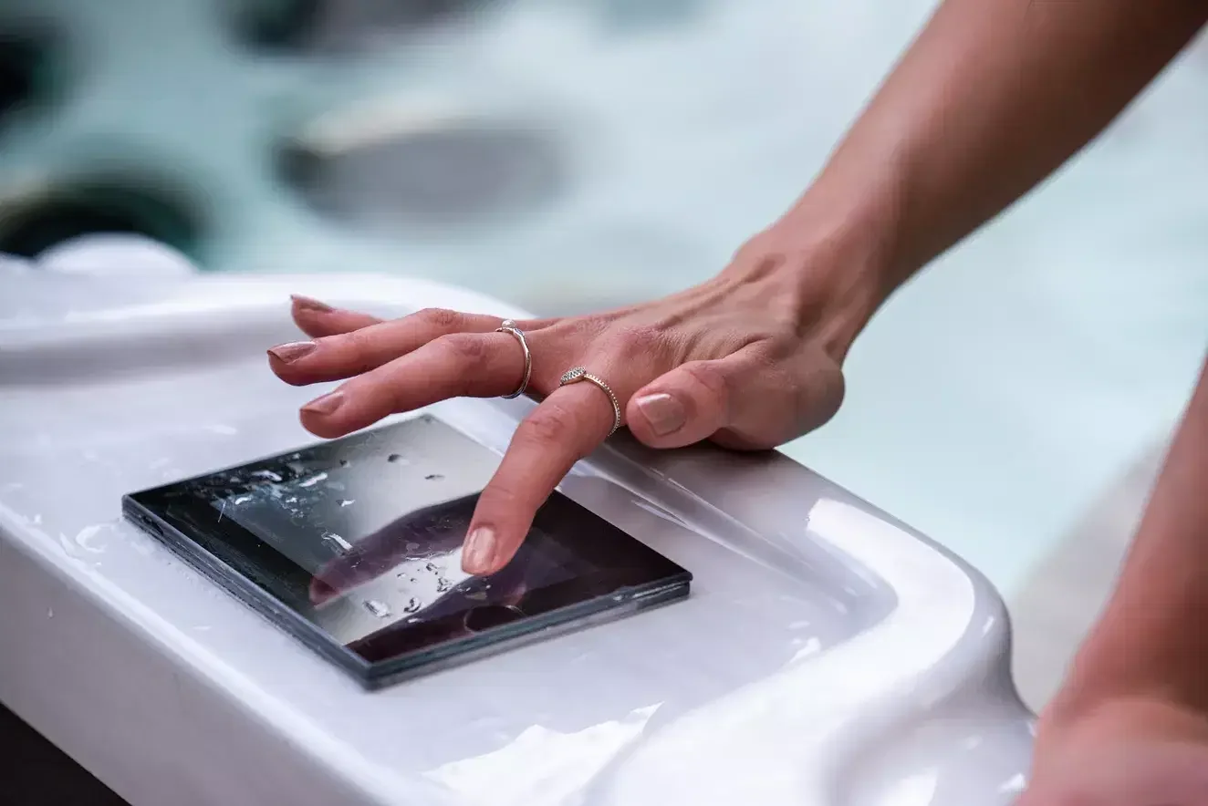 Person touching screen of a smartphone with water droplets on it, placed on a white surface.