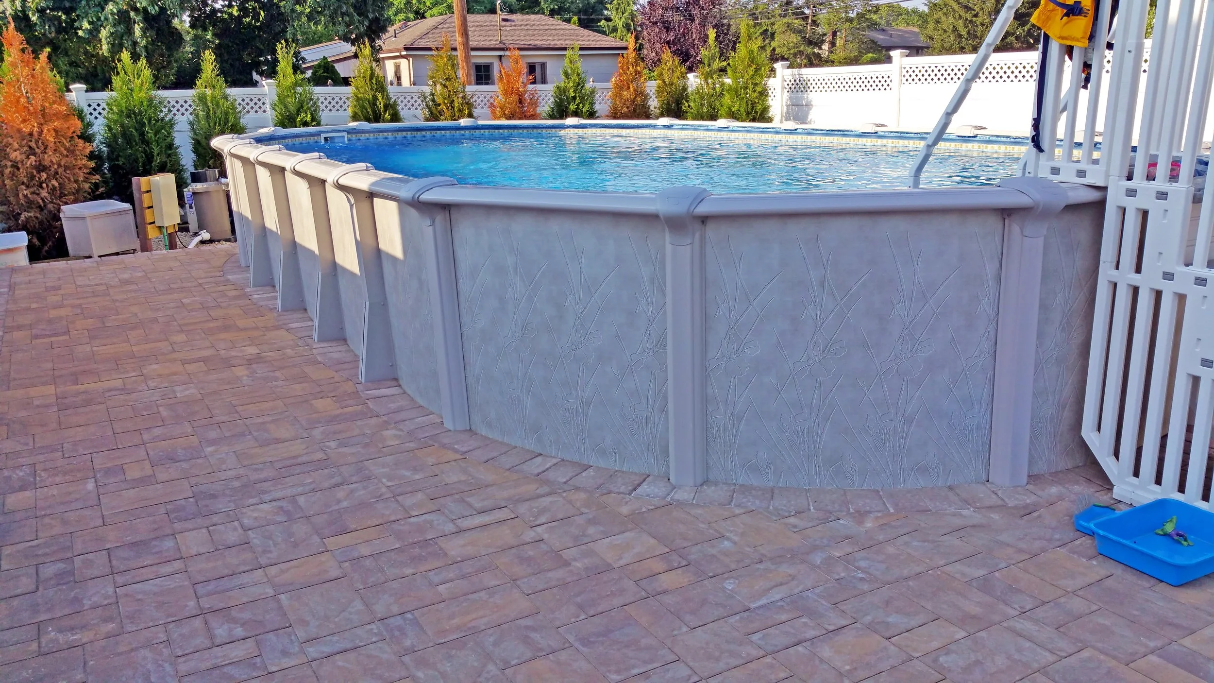 Above-ground swimming pool with a textured gray exterior, situated on a brick patio, with a view of trees, shrubs, and a white fence in the background.