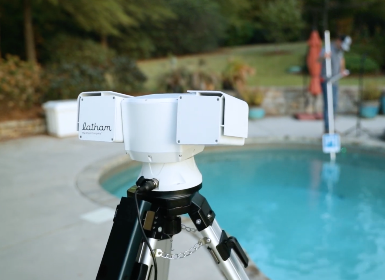 A Latham pool robot on a tripod near a backyard swimming pool, with two people standing and reading notes in the background.