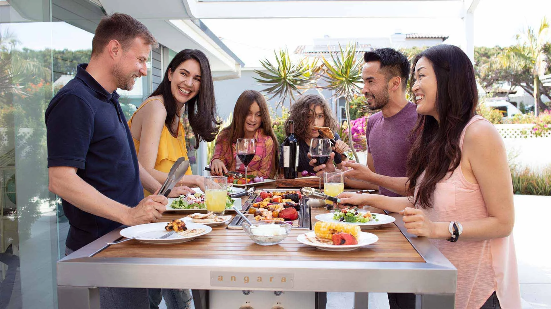 A group of seven people, including children and adults, enjoying a barbecue and dining outdoors on a patio with plants and flowers in the background.