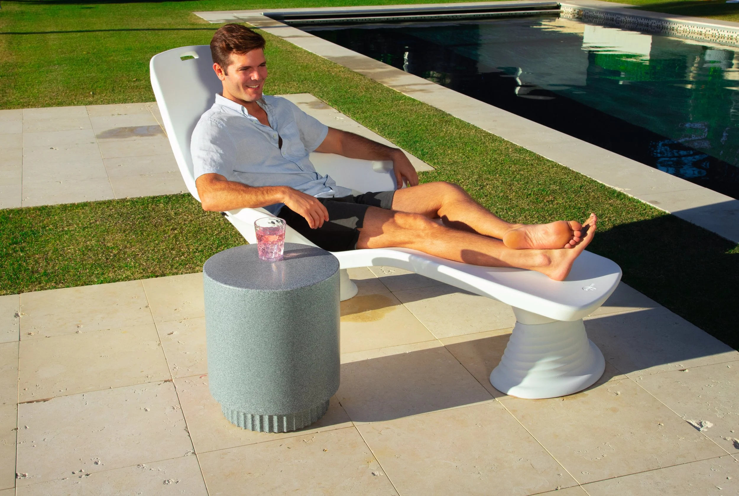 A man relaxing on a white lounge chair by a swimming pool, with a small round gray table beside him holding a glass of pink drink.
