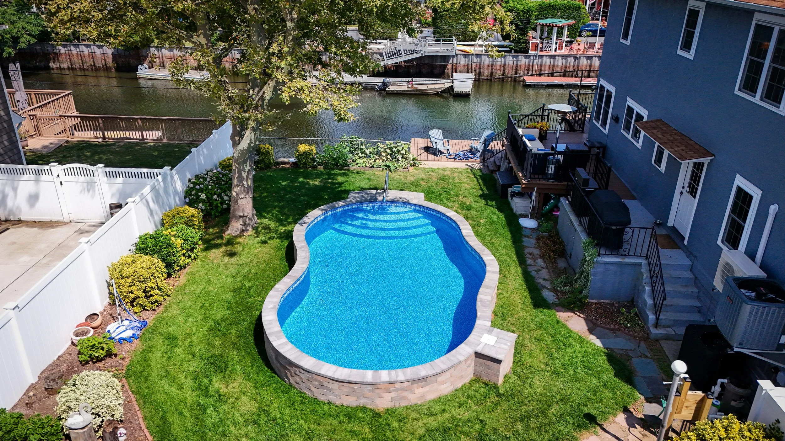 A backyard with a kidney-shaped swimming pool, green grass, a large tree, and a fence. There is a deck with chairs and a barbecue, and a river with boats and a bridge in the background.
