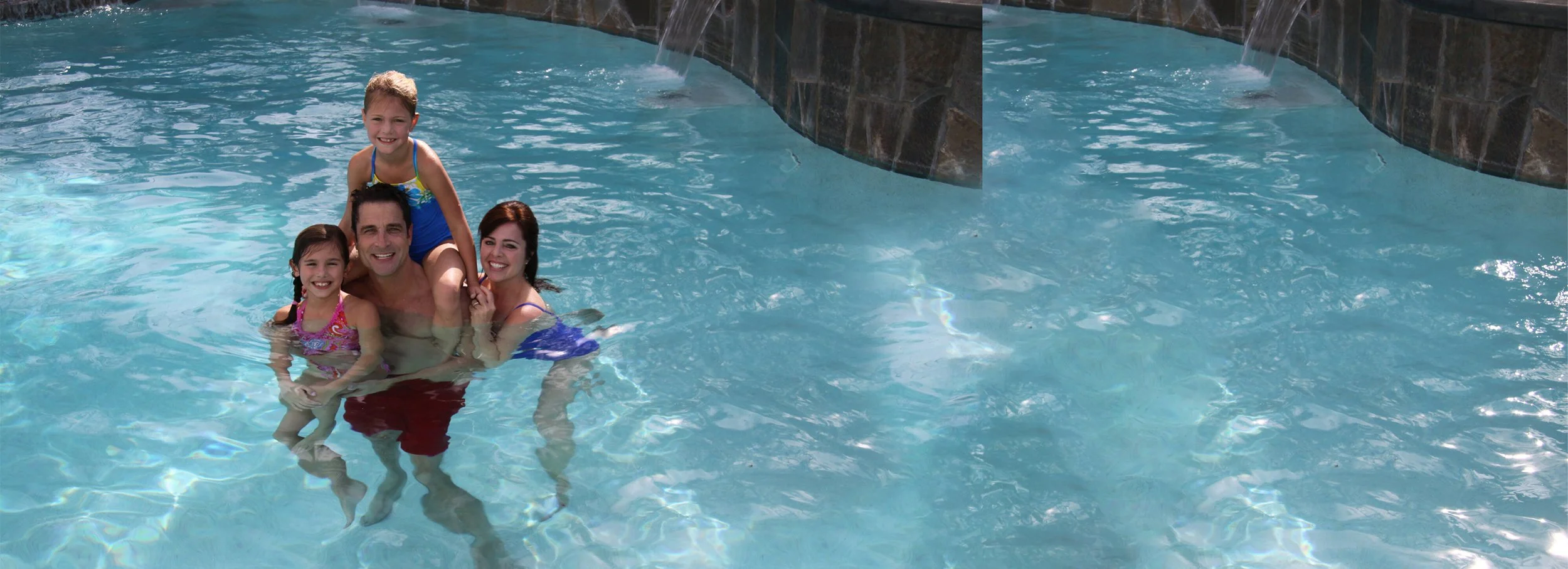 A family of four enjoying time in a swimming pool, with a waterfall feature on a stone wall in the background