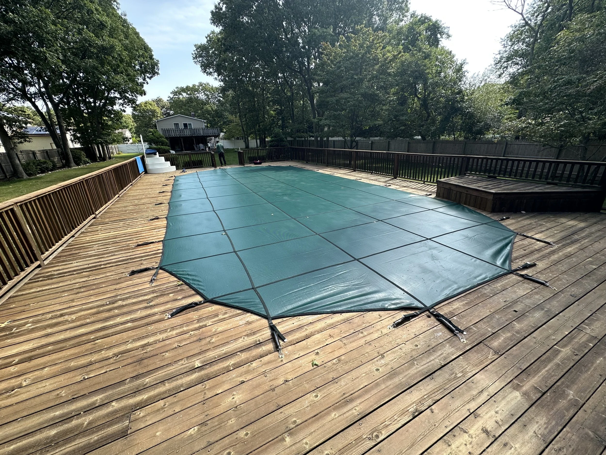 A secured covered swimming pool on a wooden deck, surrounded by a backyard with trees and a fence.