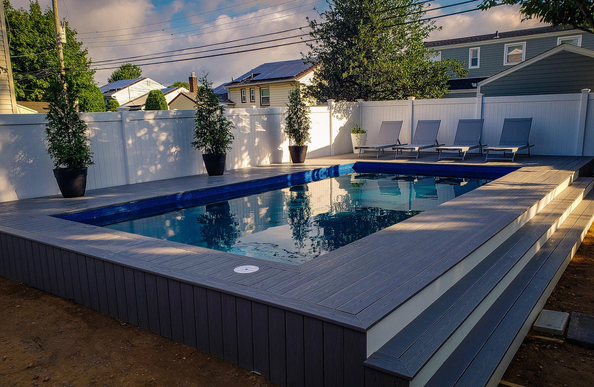 A backyard pool with a wooden deck, surrounded by a white privacy fence, with four lounge chairs and potted plants, under a partly cloudy sky.