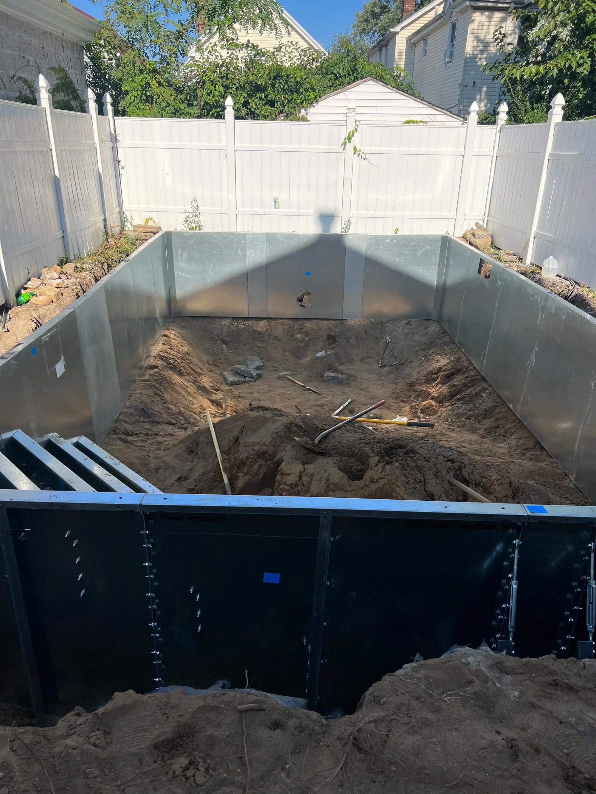 In-ground pool construction site with metal framing and dirt, surrounded by white privacy fence and residential homes.