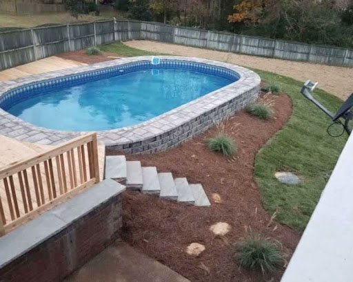 Backyard swimming pool with stone edging, small concrete stairs, and landscaped garden with grass and plants, enclosed by a wooden fence.