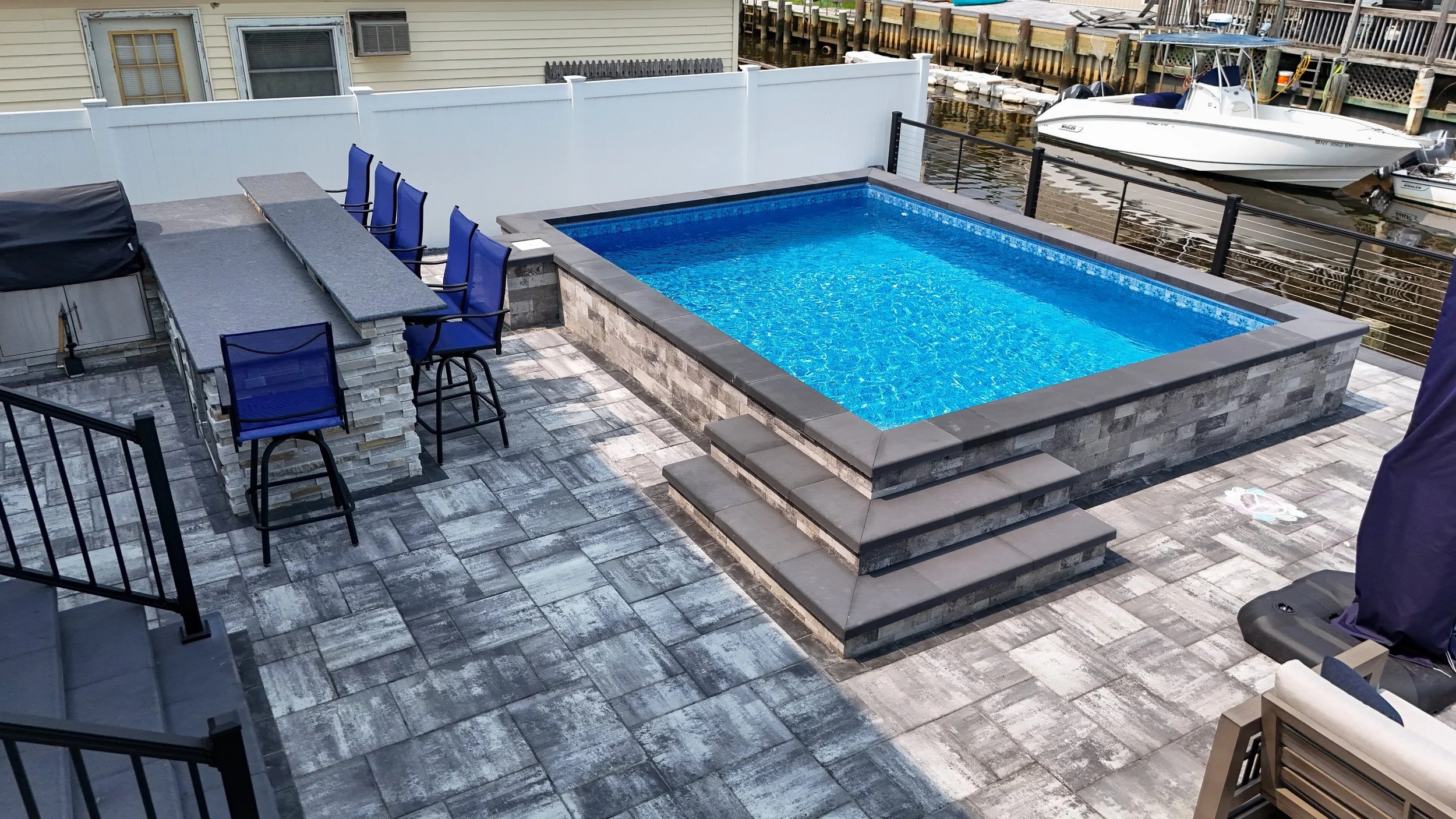 A backyard patio with a small rectangular swimming pool, elevated on a brick and stone platform, facing a waterfront with boats docked, a white fence, and a neighboring house with a window and an air conditioning unit.