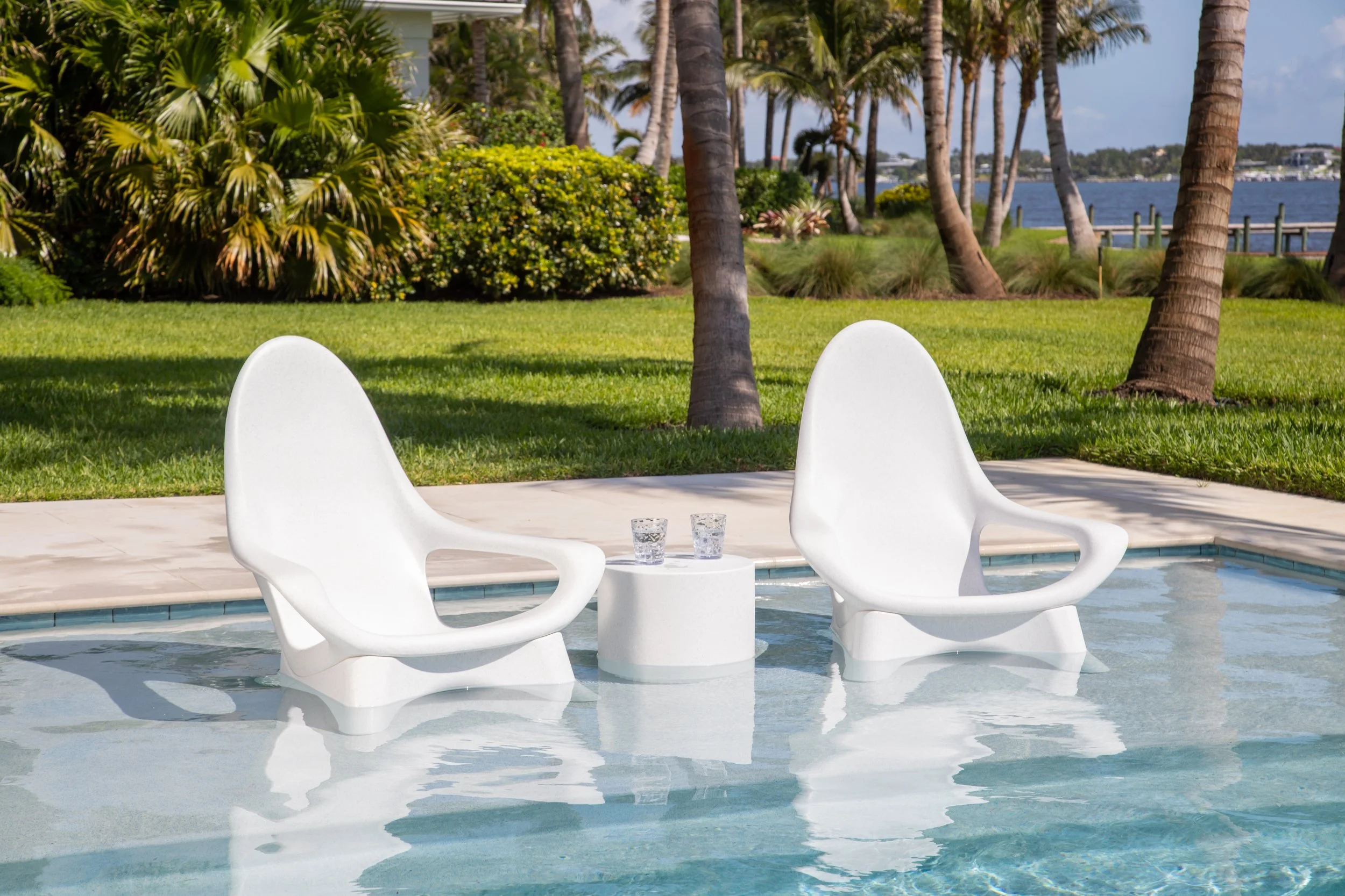 Two white modern lounge chairs in a pool with a small white table between them, holding two glasses of water. In the background, there are green lawns, bushes, and palm trees near a body of water.