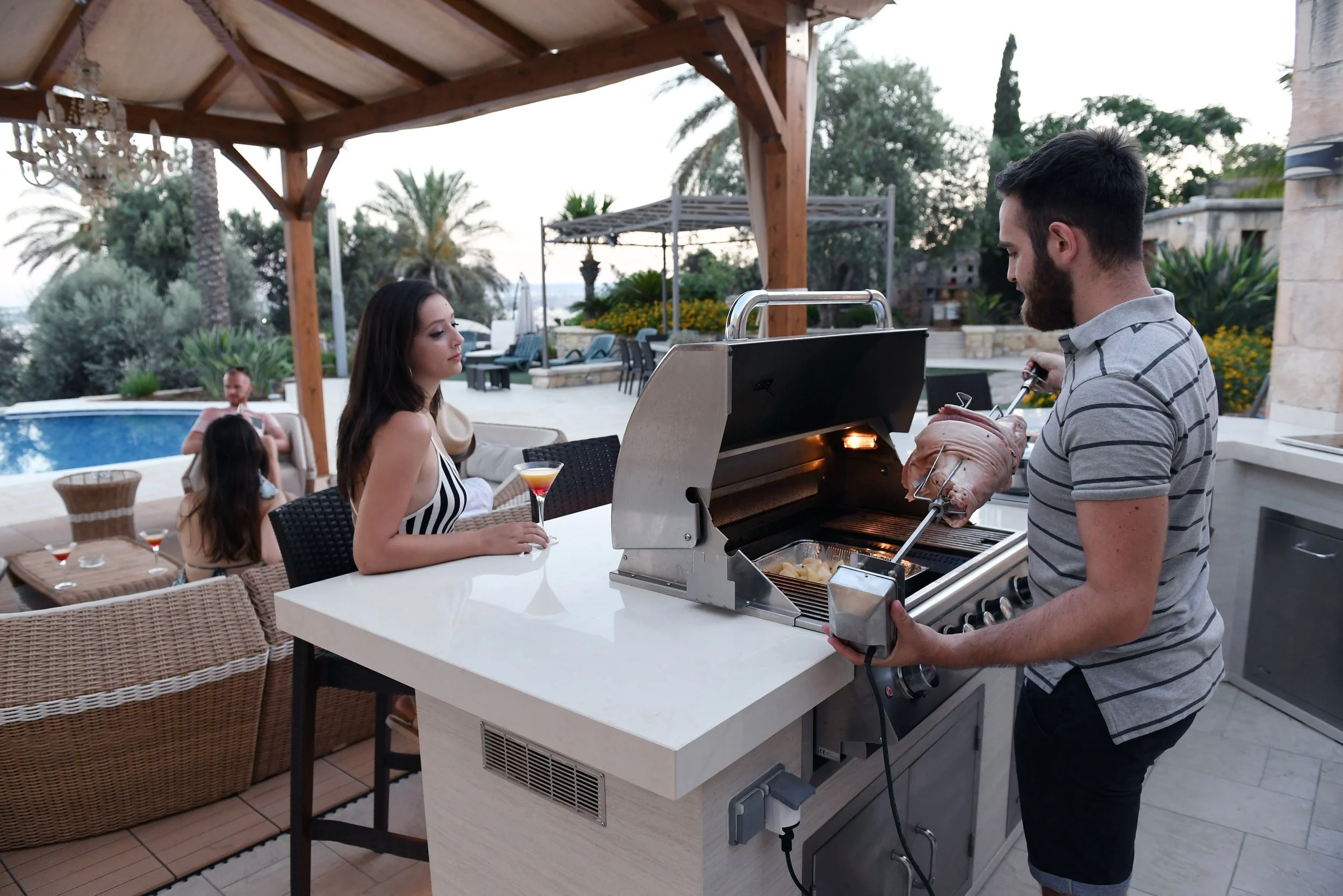 Man grilling meat on an outdoor barbecue with three women nearby, one holding a cocktail, in a backyard with pool and palm trees.