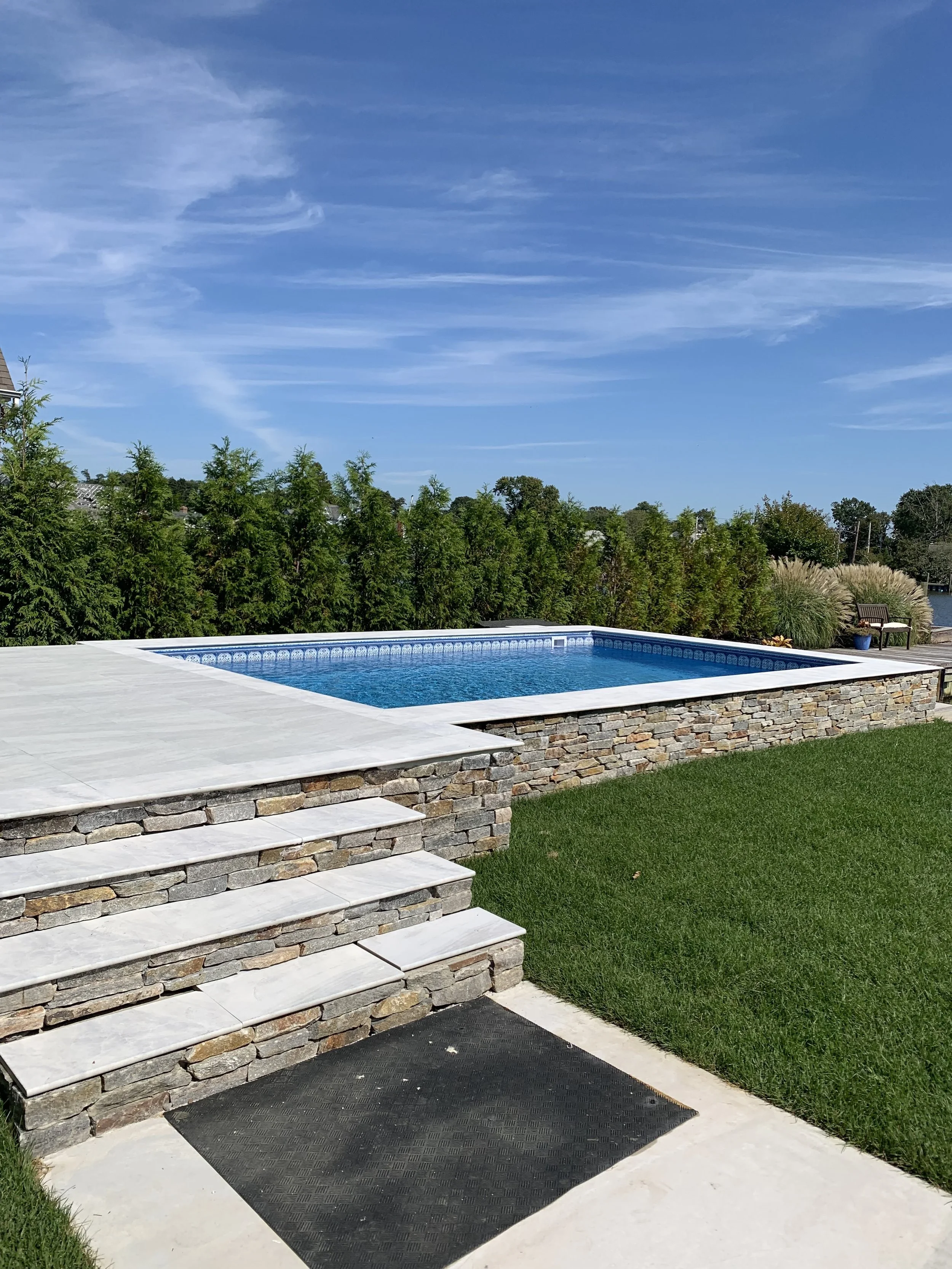 Backyard pool with stone patio stairs, surrounded by a grassy lawn, with trees and a blue sky in the background.