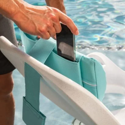 A person placing a smartphone into a zippered pocket of a teal waterproof bag next to a swimming pool.