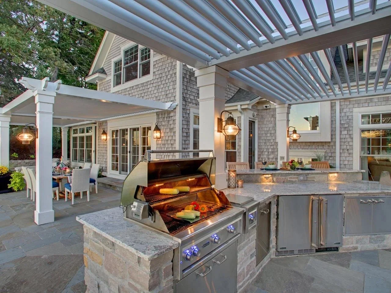 An outdoor patio area with a built-in grill cooking vegetables, a dining table with chairs, and decorative lamps, behind a house with shingle siding and large windows.