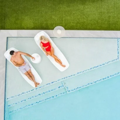 A man and woman in swimsuits lounging by a pool on pool chairs, with a poolside table and the pool's water visible.