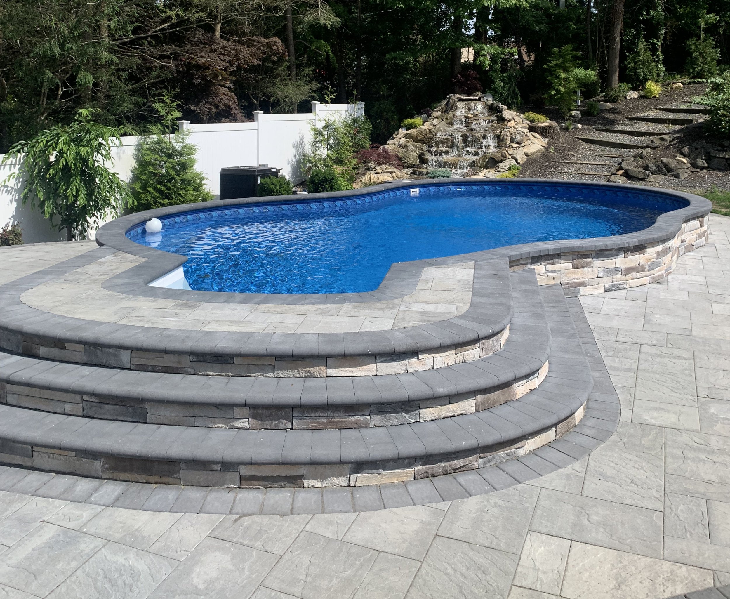 Backyard with a kidney-shaped swimming pool, stone patio, steps leading into the pool, outdoor furniture, and an American flag, surrounded by landscaped garden and trees.