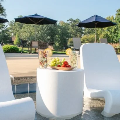 Outdoor poolside area with white modern lounge chairs, a white side table with a bowl of green grapes and strawberries, and two large navy umbrellas providing shade under a clear sky.