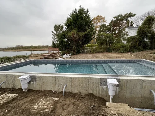 Under construction view of an in-ground swimming pool with unfinished concrete surround, pool equipment, and surrounding landscaping with trees and a wooden dock in the background.