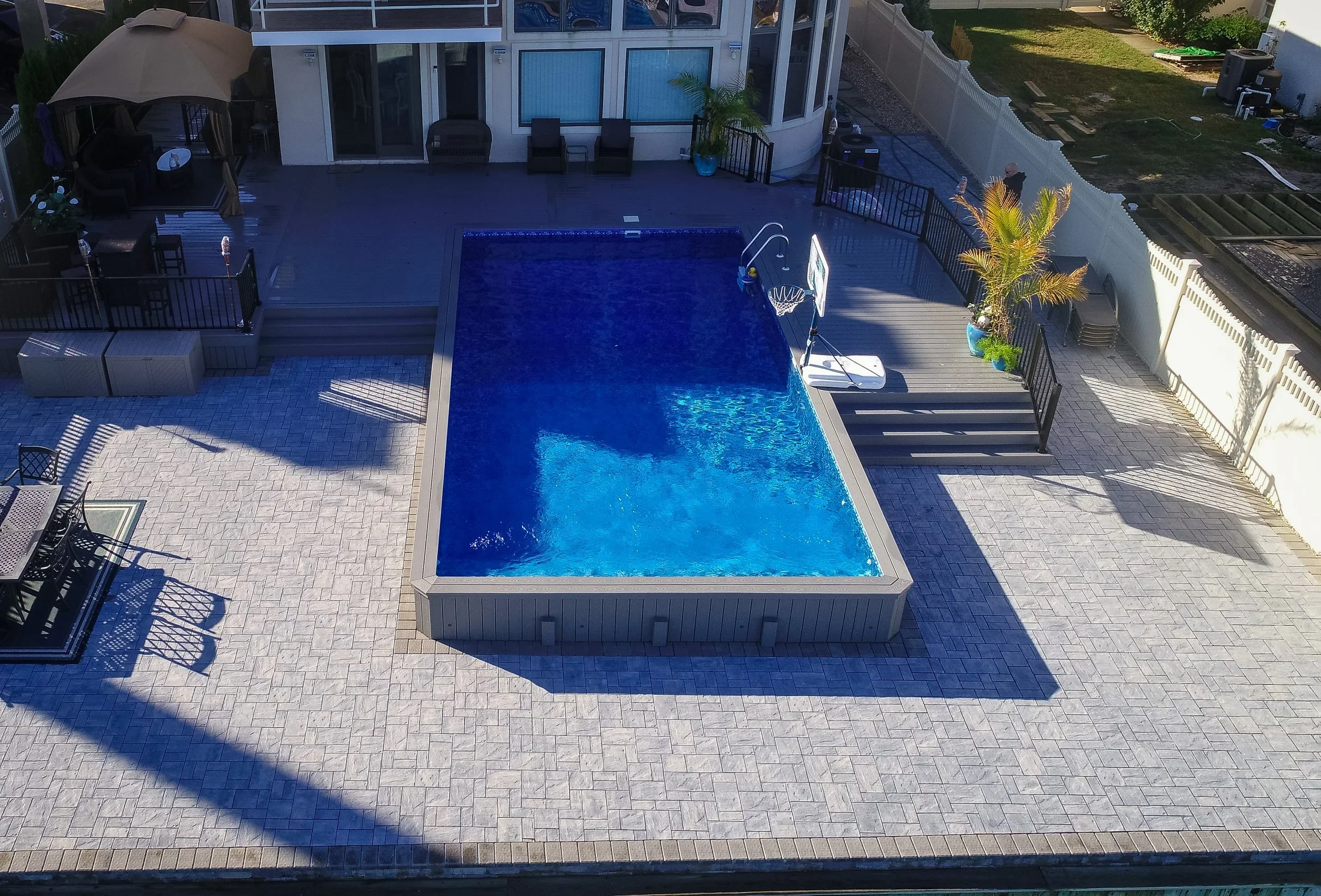 A backyard with a rectangular swimming pool, patio furniture, stairs leading to a house, and a basketball hoop near the pool.
