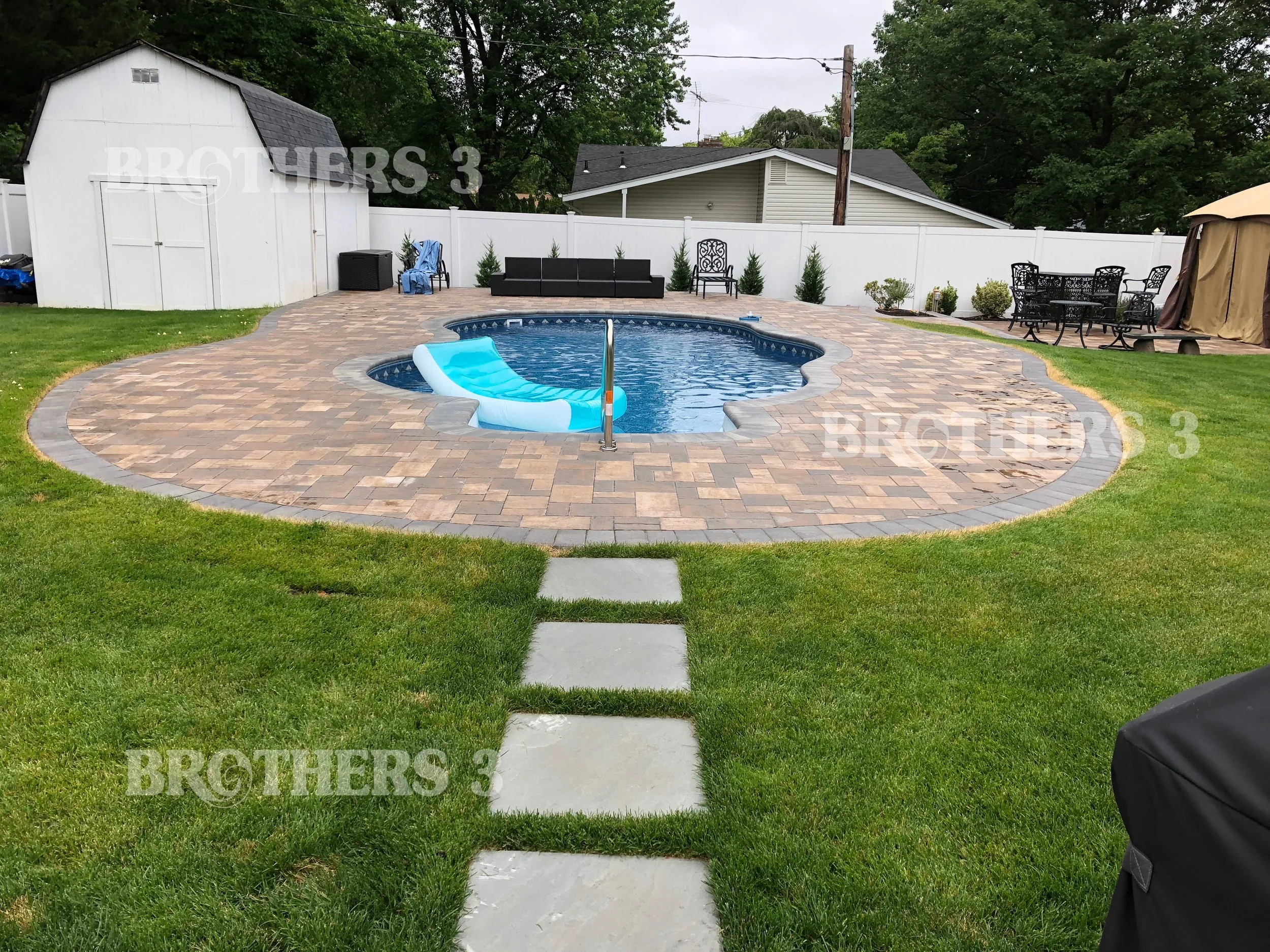 Backyard with in-ground pool surrounded by a brick and stone patio, with a float in the pool, a white shed in the back, outdoor seating including a black sofa, a table with chairs, and a gazebo. Green grass surrounds the patio, and there are trees and houses in the background.
