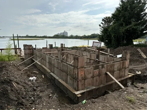 A partially built concrete foundation in a construction site by a body of water, with wooden formwork and construction materials around.
