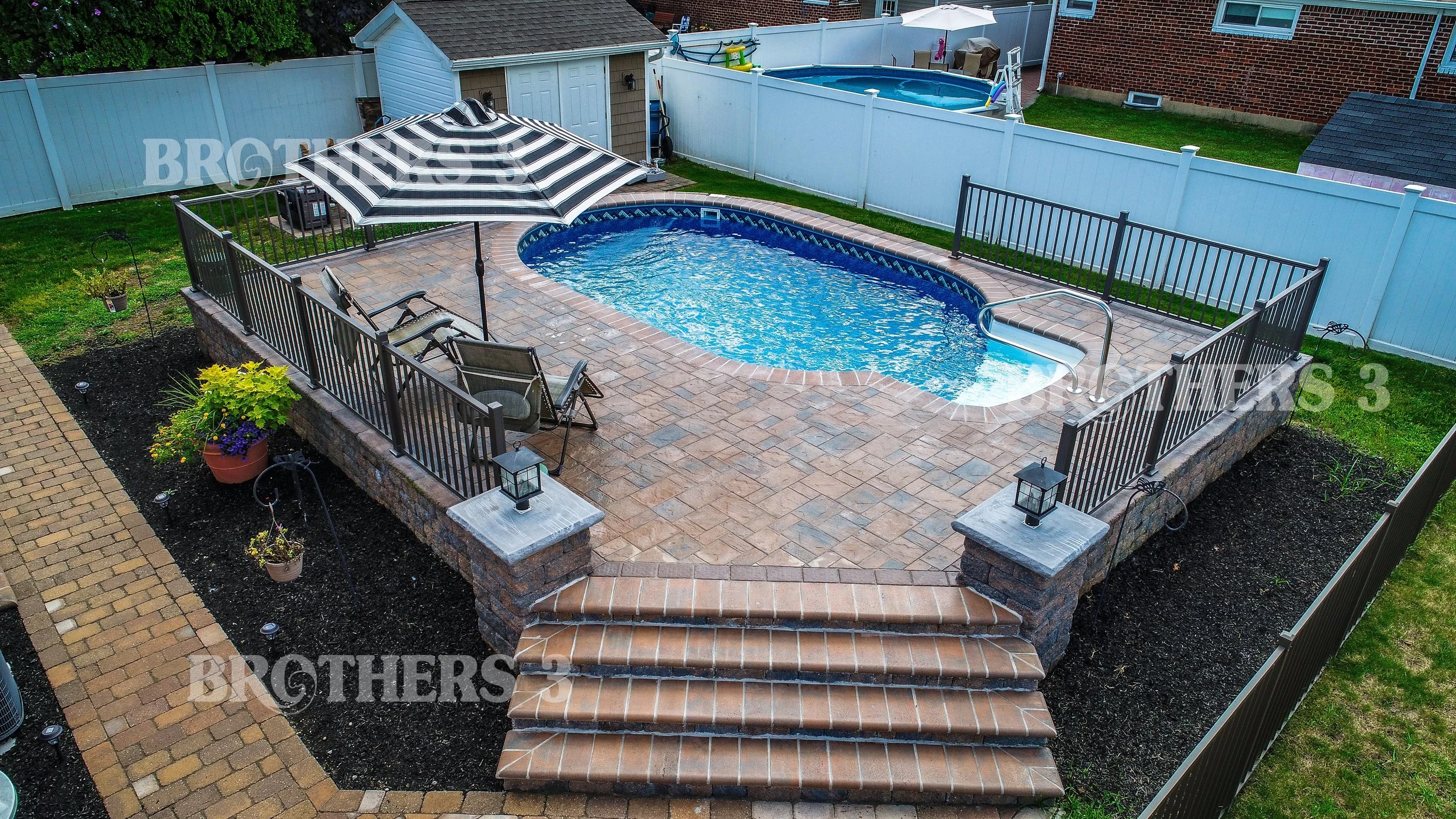 Backyard with a small in-ground swimming pool, brick patio, black metal fence, patio chairs, striped umbrella, steps leading up to the patio, small lawn area, potted plants, garden lights, a shed, a trampoline, and a brick house with windows in the background.